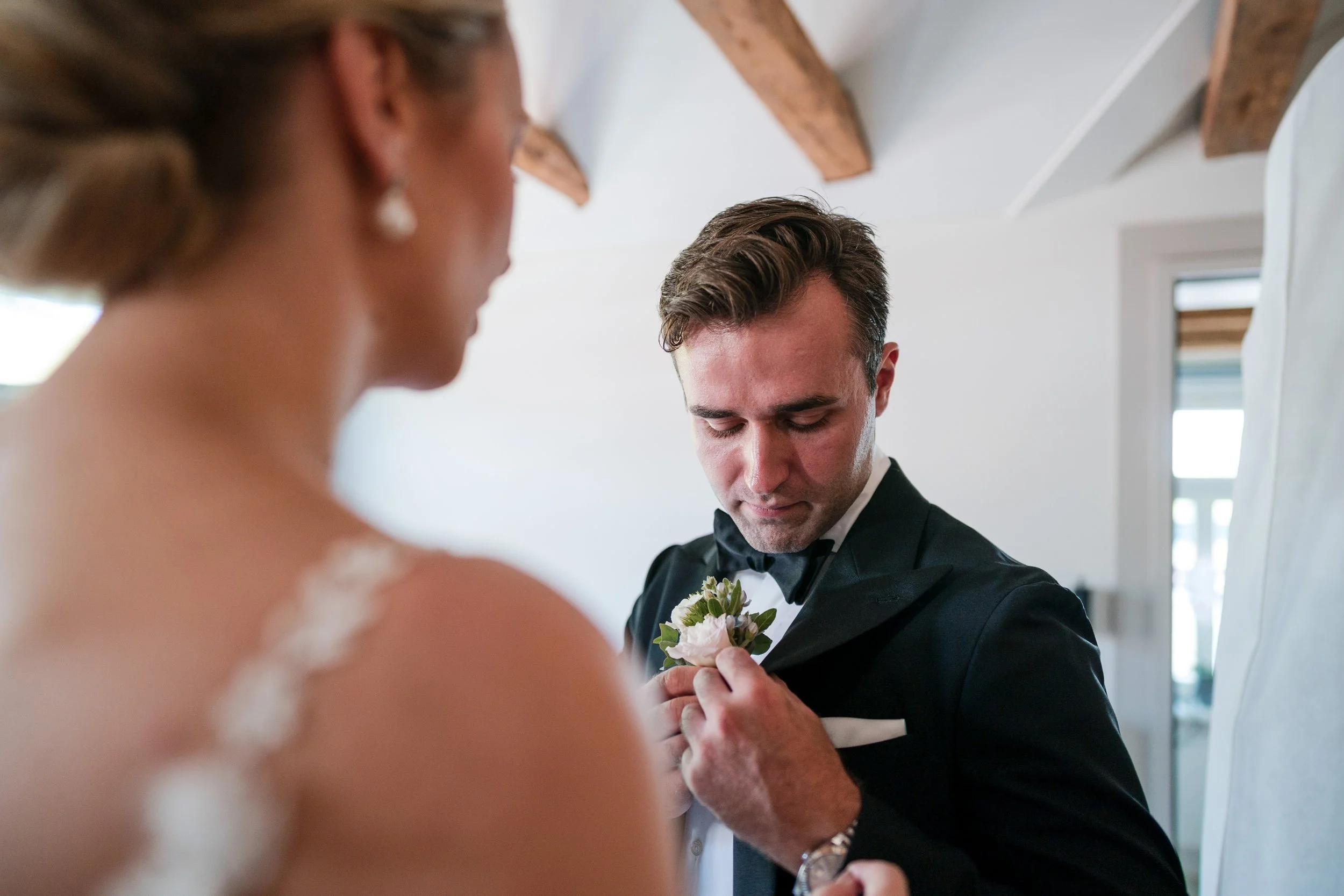 Ein Hochzeitsfotografie, zeigt einen Mann in einem schwarzen Anzug und Fliege, der eine Weste trägt, der an seinem Brustknopf eine kleine weiße Blume befestigt, während eine Frau im Vordergrund unscharf ist. Die Szene findet wahrscheinlich vor einer 
