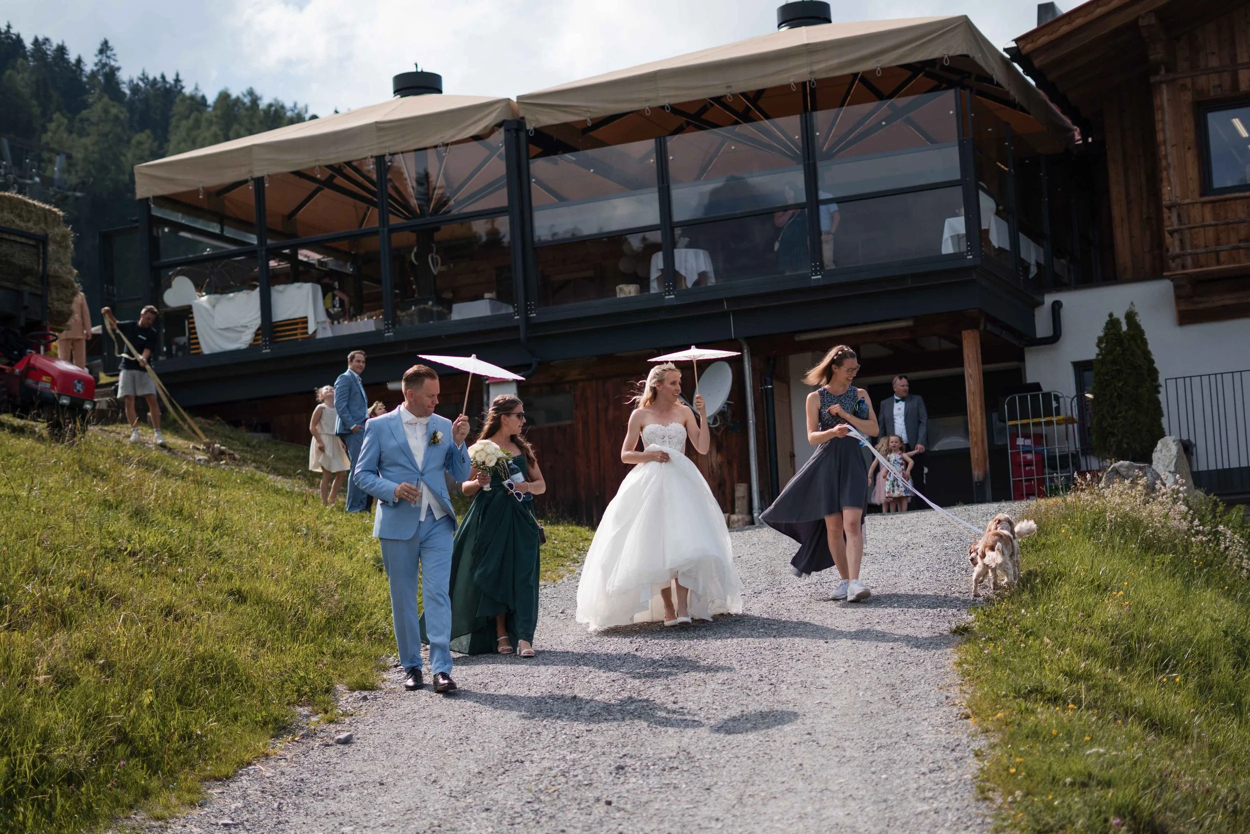 Hochzeit mit Brautpaar und Gästen, die draußen vor einem Chalet spazieren gehen, mit einem Hund, im Grünen bei sonnigem Wetter.