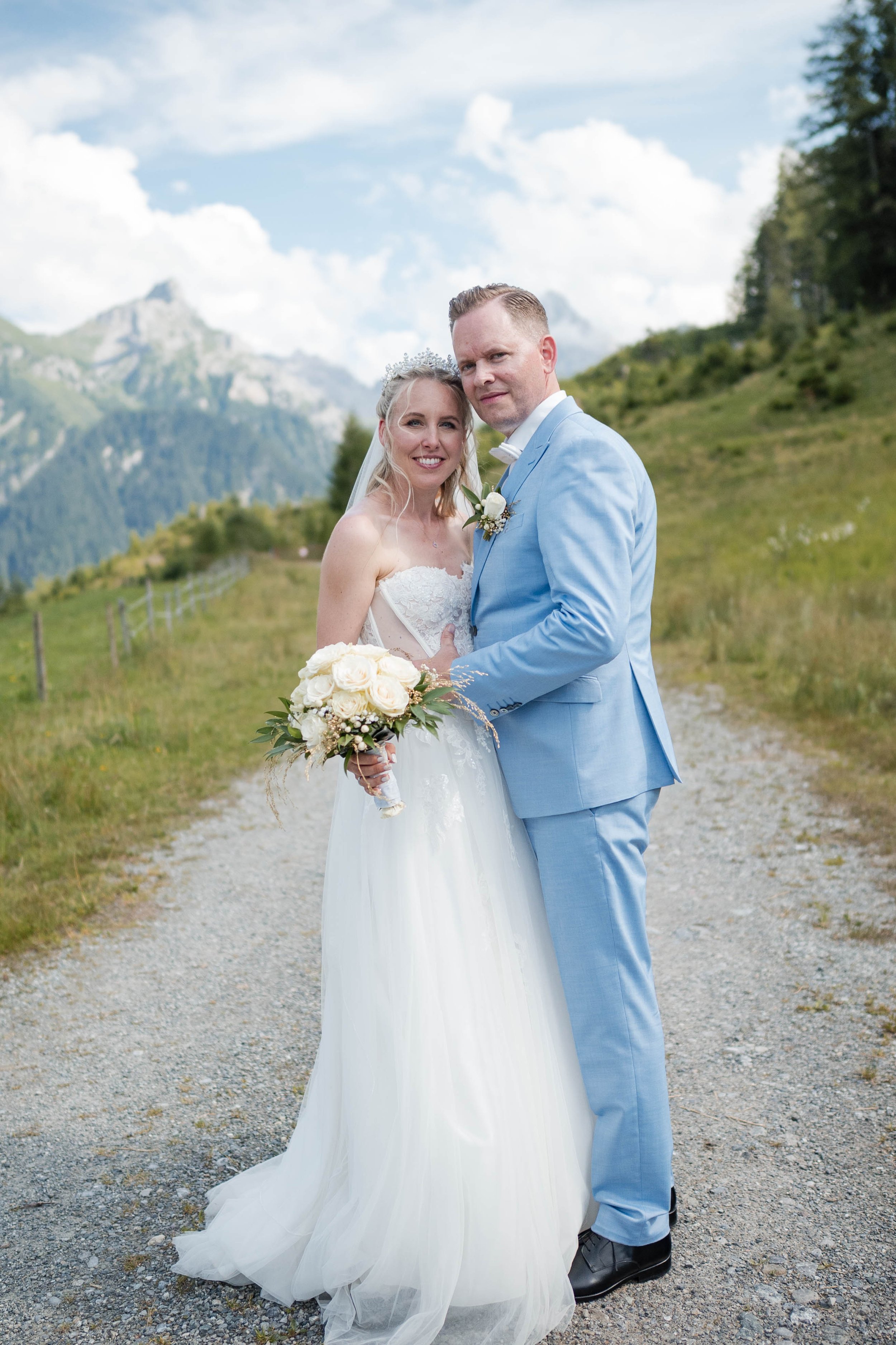 Ein Brautpaar auf einer Waldweg in den Bergen bei ihrer Hochzeit, das Mädchen trägt ein weißes Brautkleid und der Junge einen hellblauen Anzug, sie halten eine Brautstrauß, der Himmel ist bewölkt.