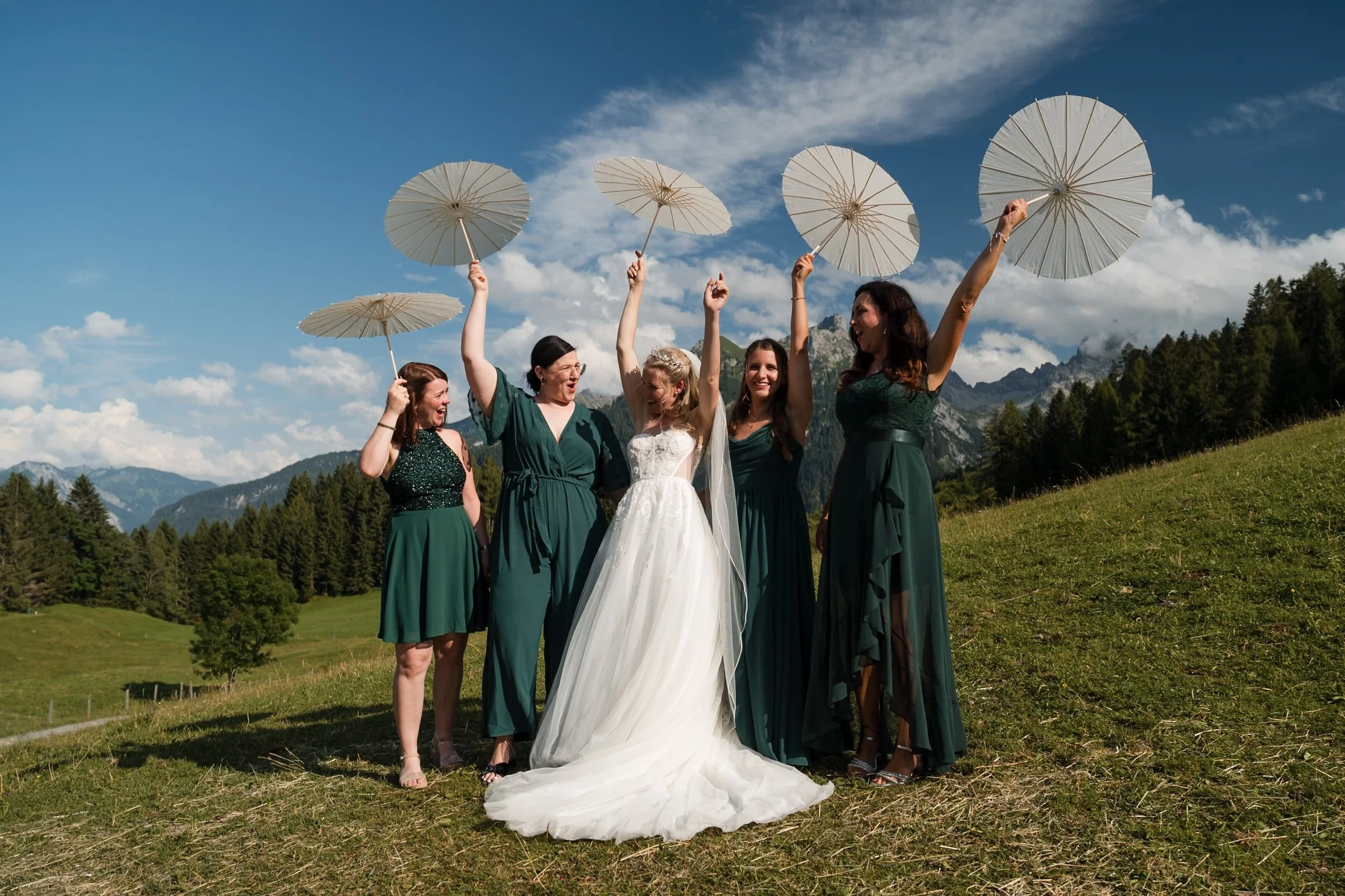Eine Braut und vier Frauen in grünen Kleidern feiern im Freien, halten weiße Sonnenschirme, vor Bergen und blauen Himmel.
