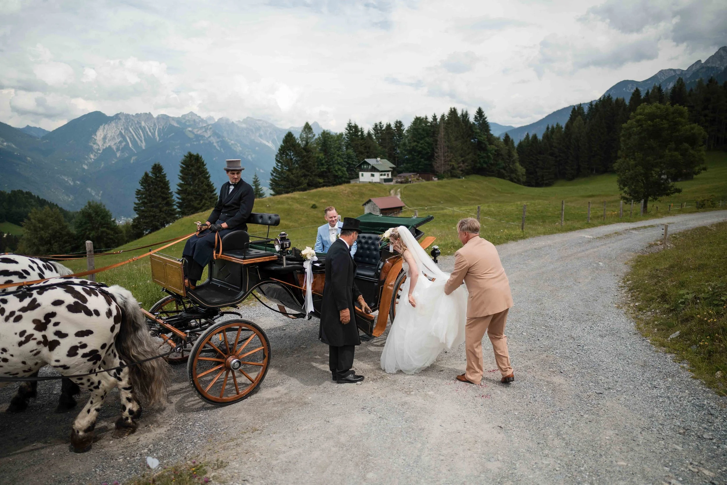 Braut und Bräutigam steigen in eine Kutsche, während ein Kutscher sie in einer ländlichen Bergregion abfährt. Es gibt eine atemberaubende Bergkulisse im Hintergrund.