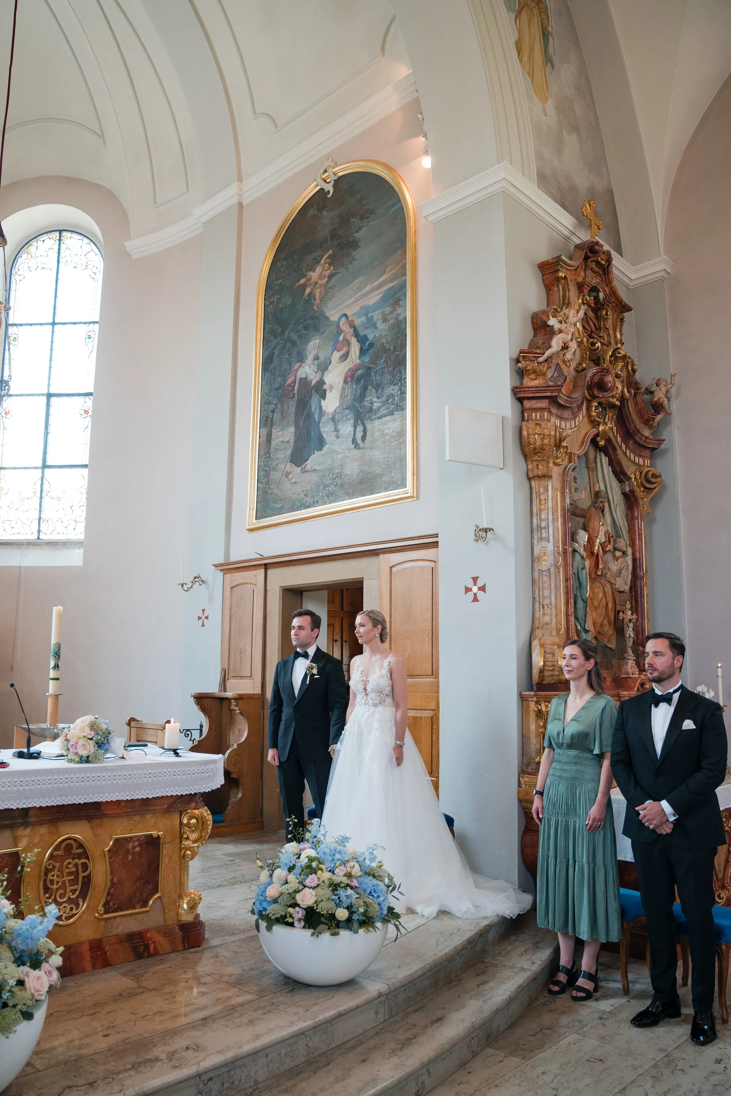 Hochzeitszeremonie in einer Kirche mit Braut, Bräutigam, zwei weiteren Personen und Altar mit Blumen und Kerzen.