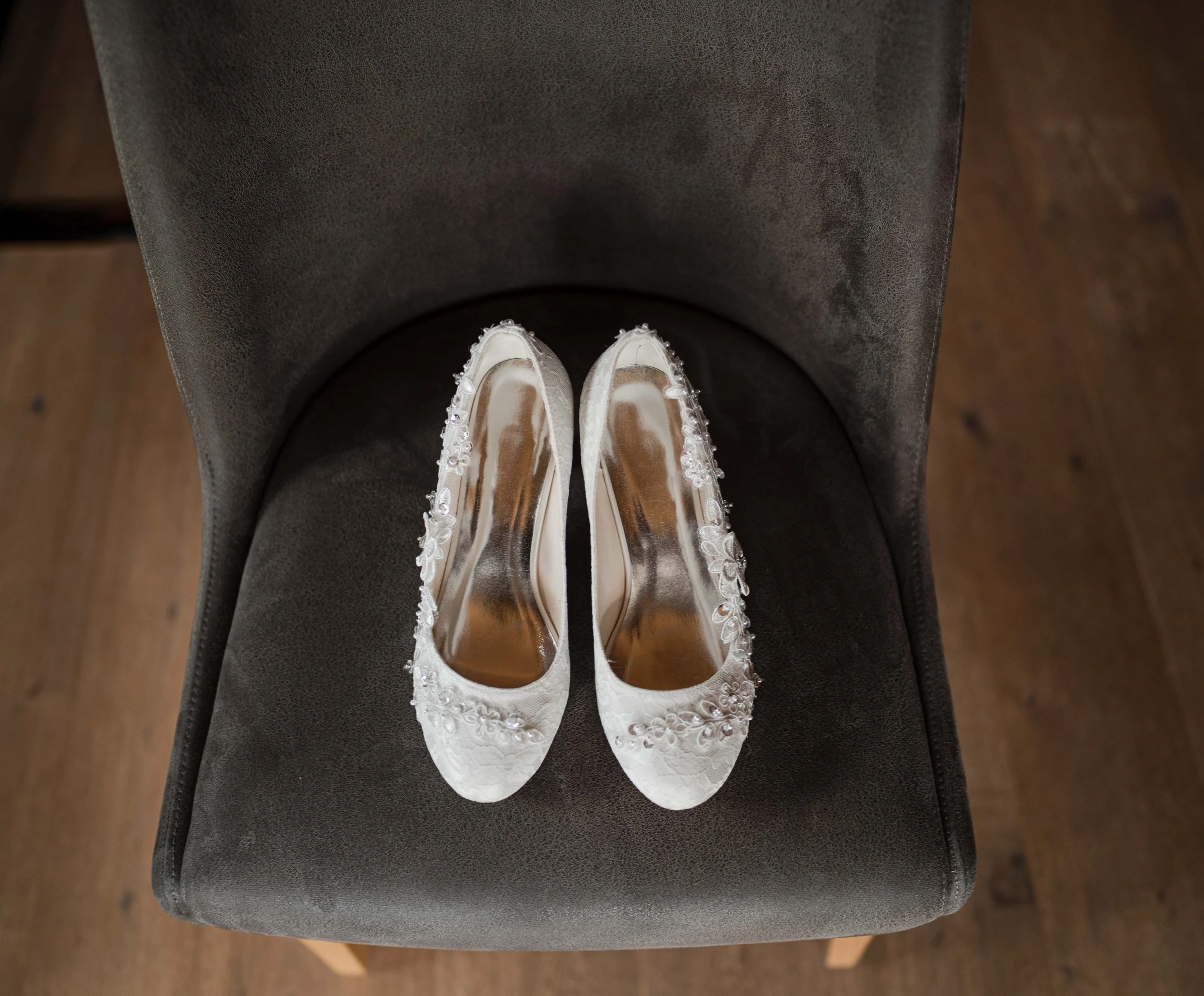 Weiße Brautschuhe mit Blumenverzierungen auf einem schwarzen Stuhl.