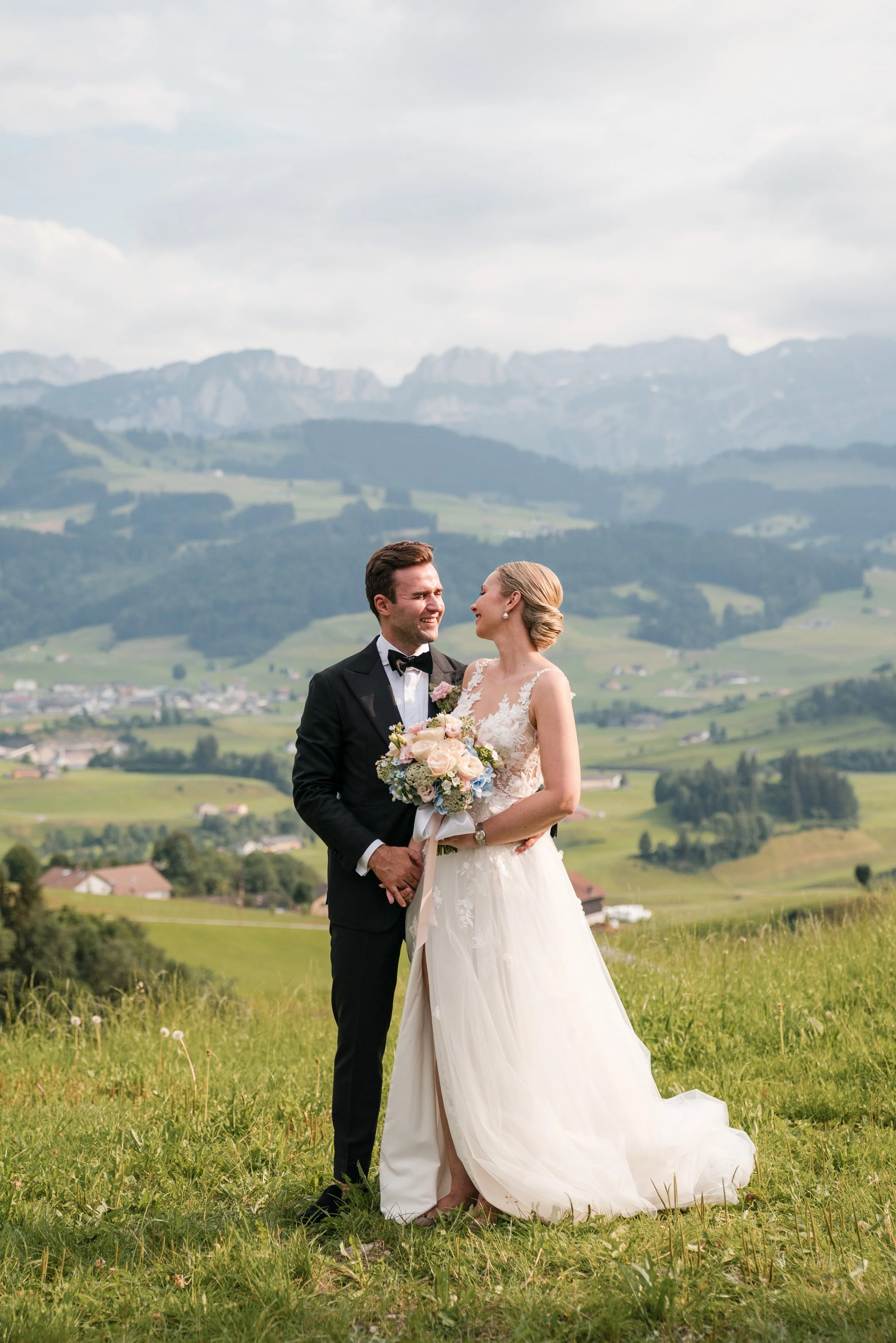 Ein Brautpaar, der Bräutigam in einem schwarzen Anzug und einer Fliege, die Braut in einem weißen Hochzeitskleid, steht auf einer grünen Wiese mit Bergen im Hintergrund, bei sonnigem Himmel.