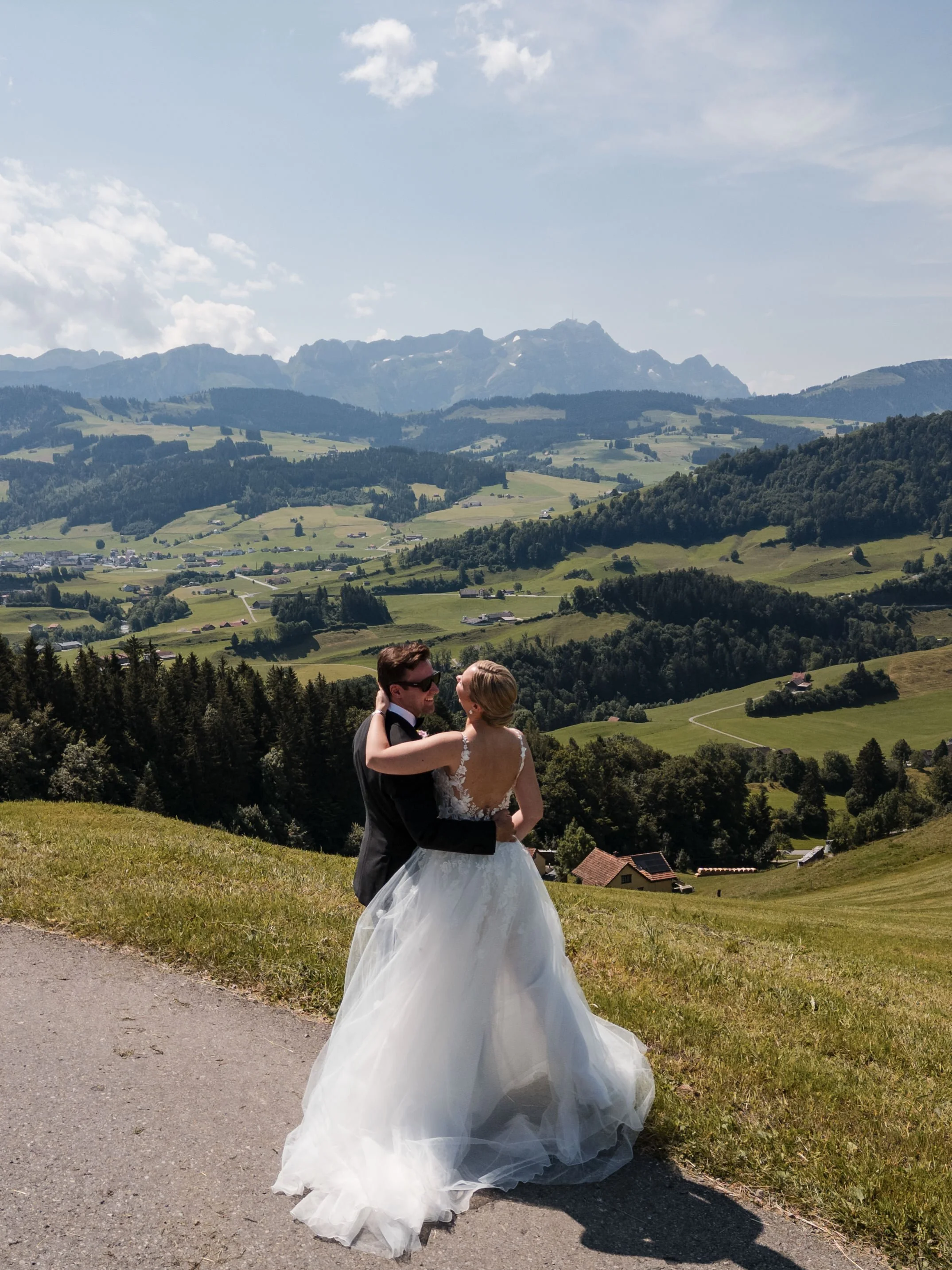 Ein Brautpaar tanzt auf einer Wiese vor einer malerischen Berglandschaft mit grünen Hügeln und fernen Bergen im Hintergrund bei schönem Wetter.