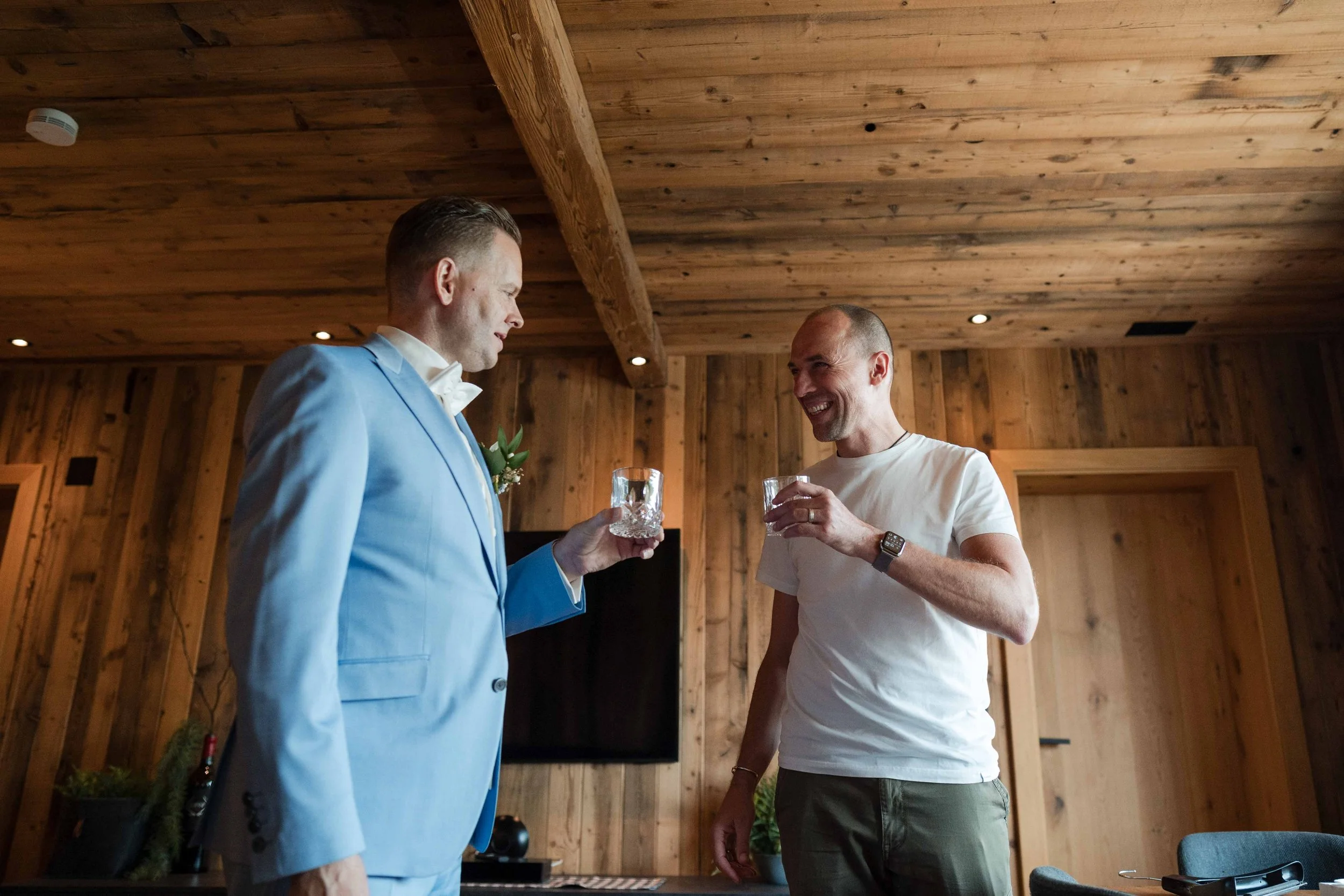 Zwei Männer stoßen sich in einem Holzraum mit Whiskey-Gläsern an, einer trägt einen hellblauen Anzug, der andere ein weißes T-Shirt und grüne Hosen, beide lachen.