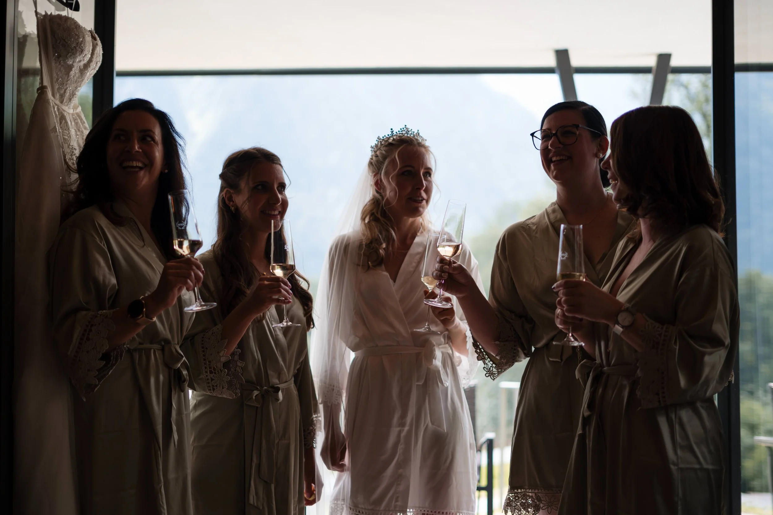 Eine Braut in weißem Kleid mit Diadem hält ein Glas Sekt, umgeben von fünf Freundinnen in einheitlichen Bademänteln, die ebenfalls Sekt trinken, in einem Raum mit großem Fenster im Hintergrund.