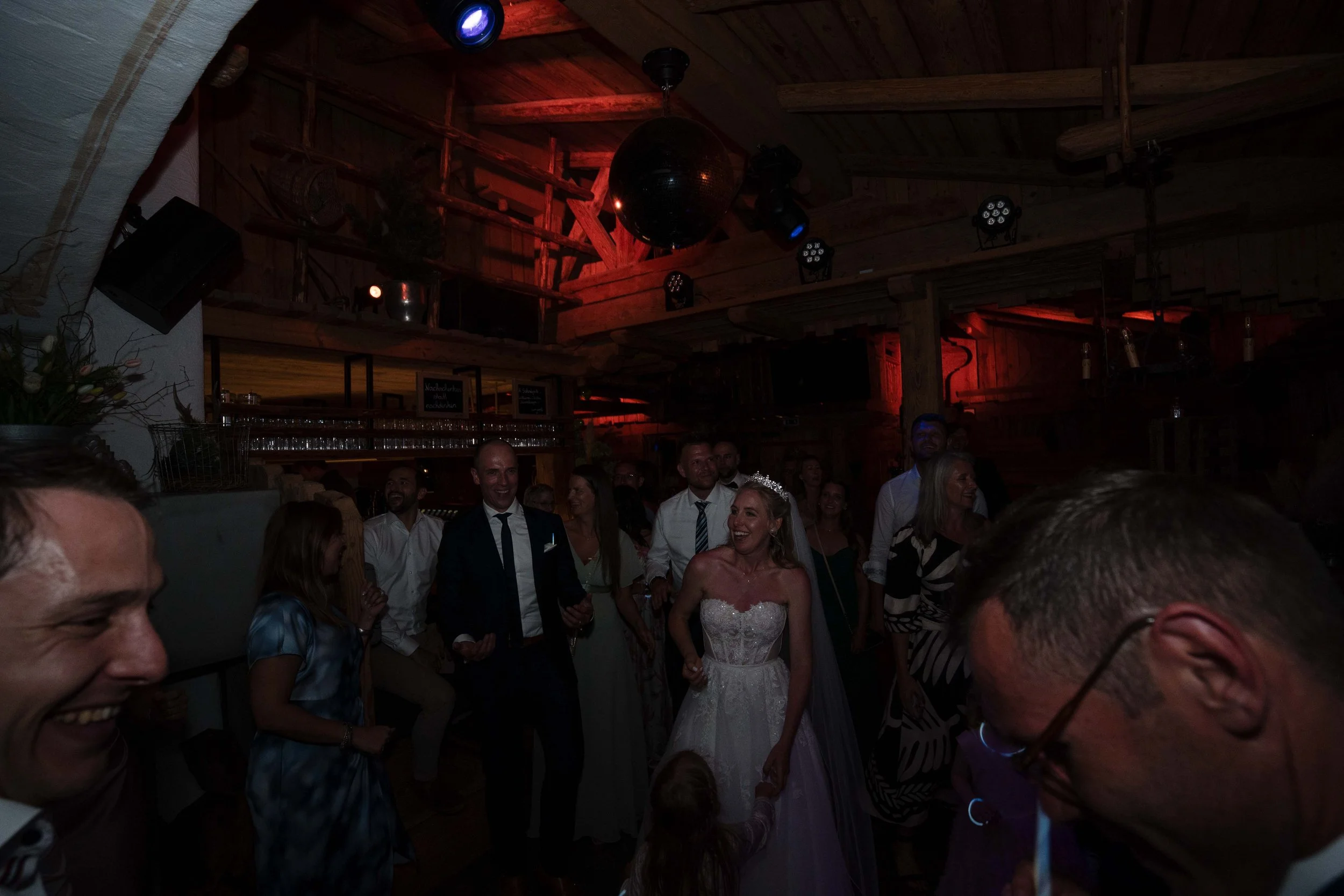 Hochzeitsparty in einem rustikalen Holzhaus mit Braut im weißen Kleid, umgeben von Gästen, bei Tanz und Feier.