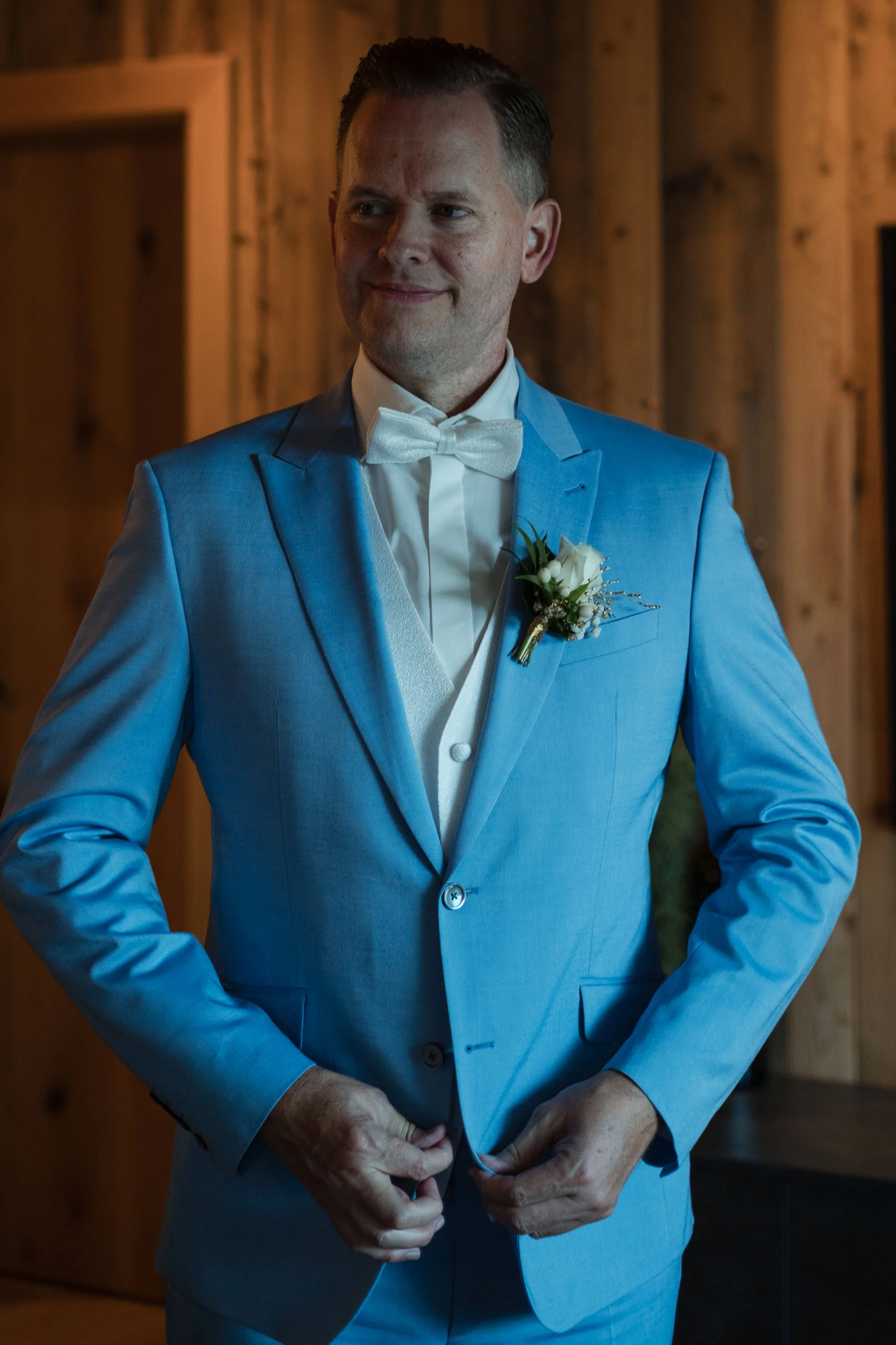 Mann in blauer Anzug mit weißem Hemd und Fliege, trägt einen weißen Blumenstrauß auf der Brust, steht in einem Raum mit Holzdekoration.