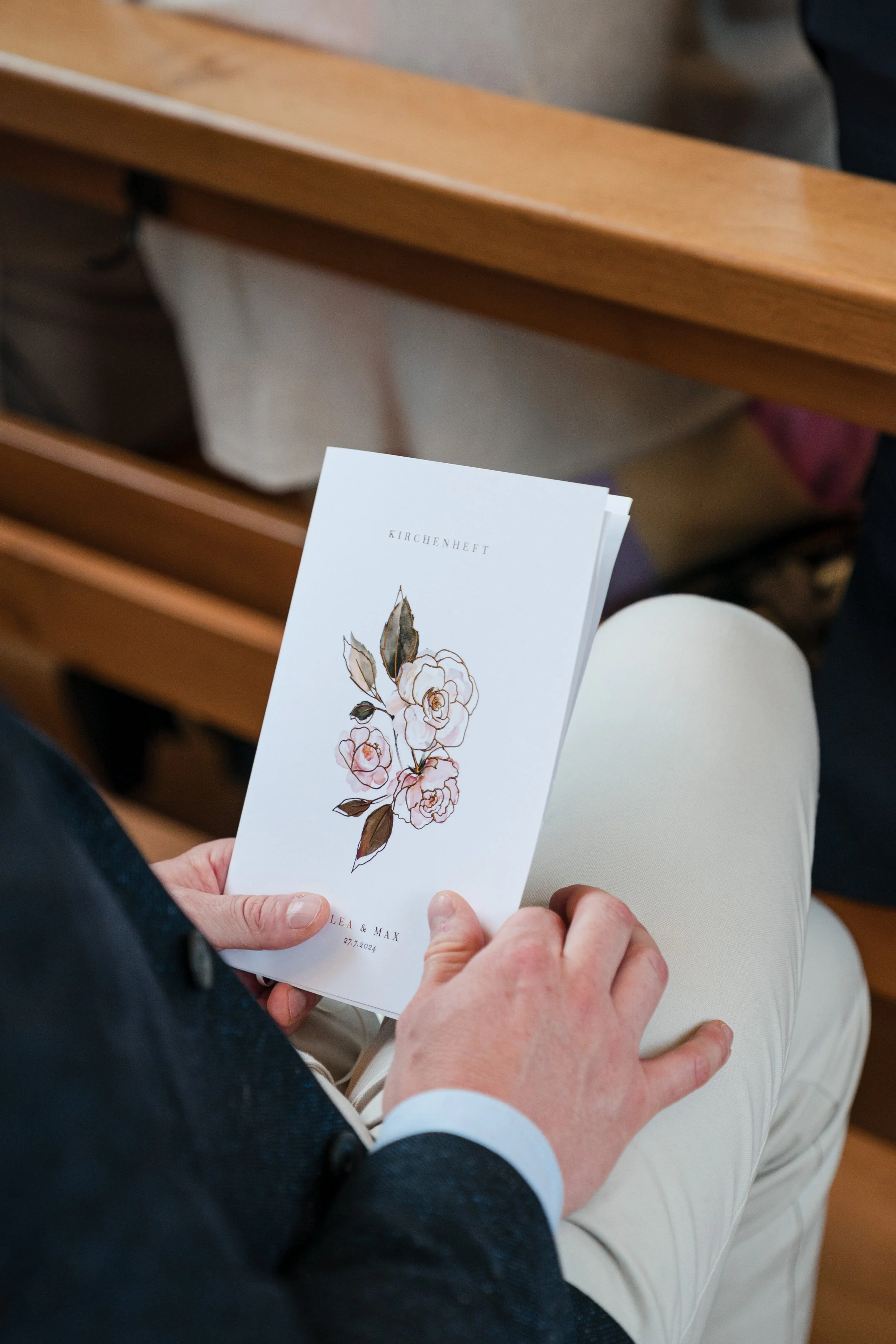 Person hält ein Kirchenheft mit einer Blumenzeichnung auf dem Schoß, sitzend auf einem Sitz in einer Kirche.