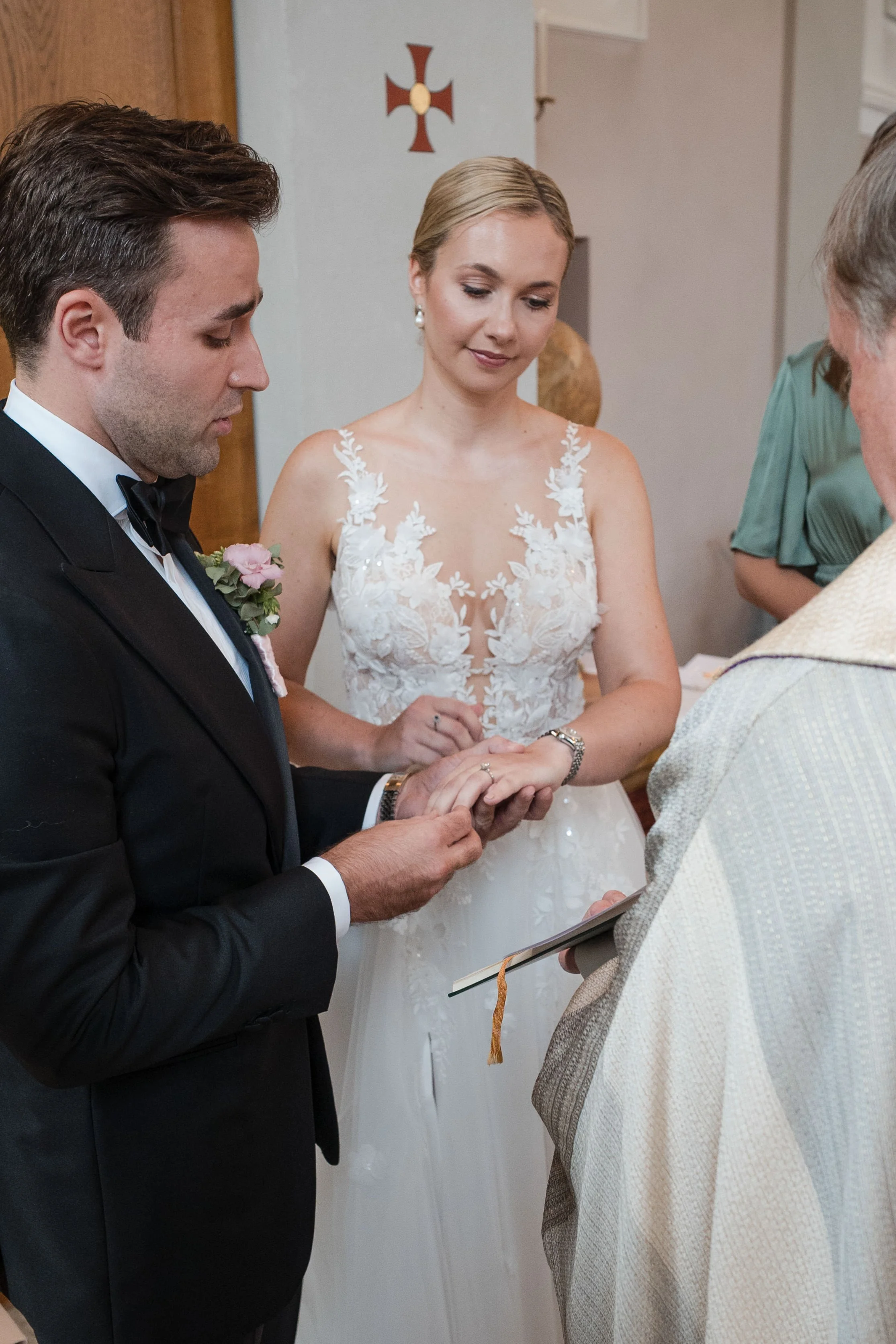 Ein Brautpaar bei der Trauungszeremonie, der Bräutigam legt den Ehering am Finger der Braut an, im Hintergrund christliches Kreuz an der Wand.