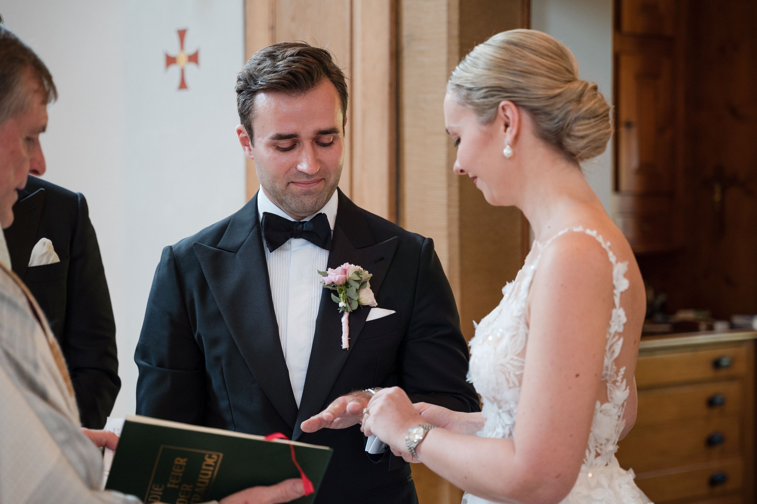 Ein Brautpaar bei der Trauung, der Bräutigam trägt einen schwarzen Anzug mit Fliege, die Braut trägt ein weißes Hochzeitskleid mit Spitze, sie tauschen Ringe.