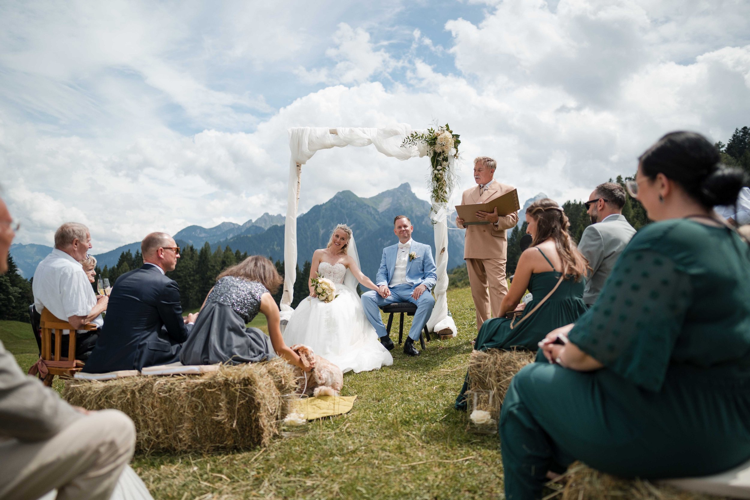 Eine Heirat im Freien vor malerischer Bergkulisse, bei der das Brautpaar unter einem weißen Blumen- und Stoffdekor einen Trauungen absolviert, umgeben von Gästen, die auf Heuballen sitzen.