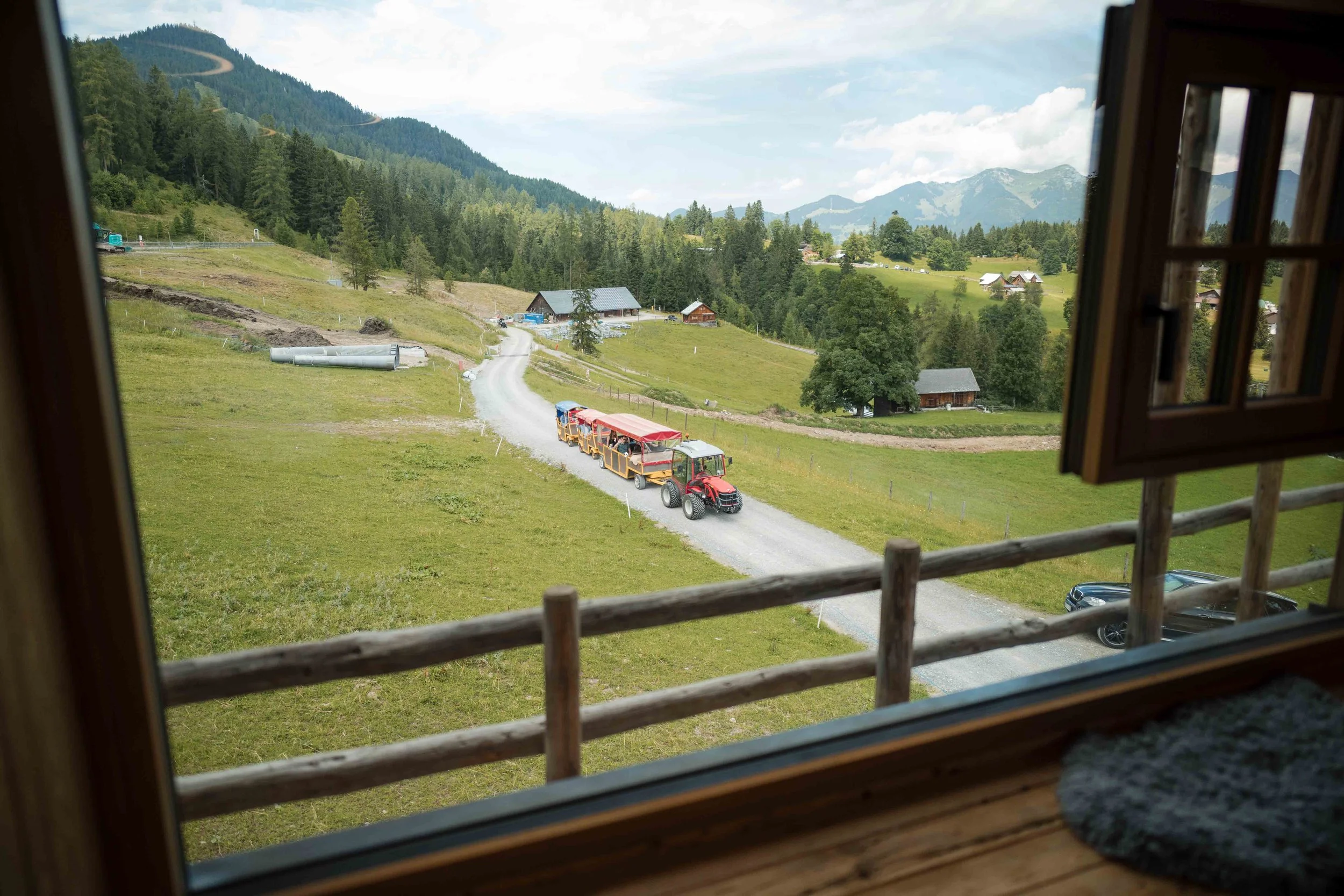 Blick aus einem Fenster auf eine ländliche Landschaft mit einem kleinen Zug, der auf einer Schotterstraße fährt, umgeben von grünen Wiesen und Hügeln, mit Bergen im Hintergrund.
