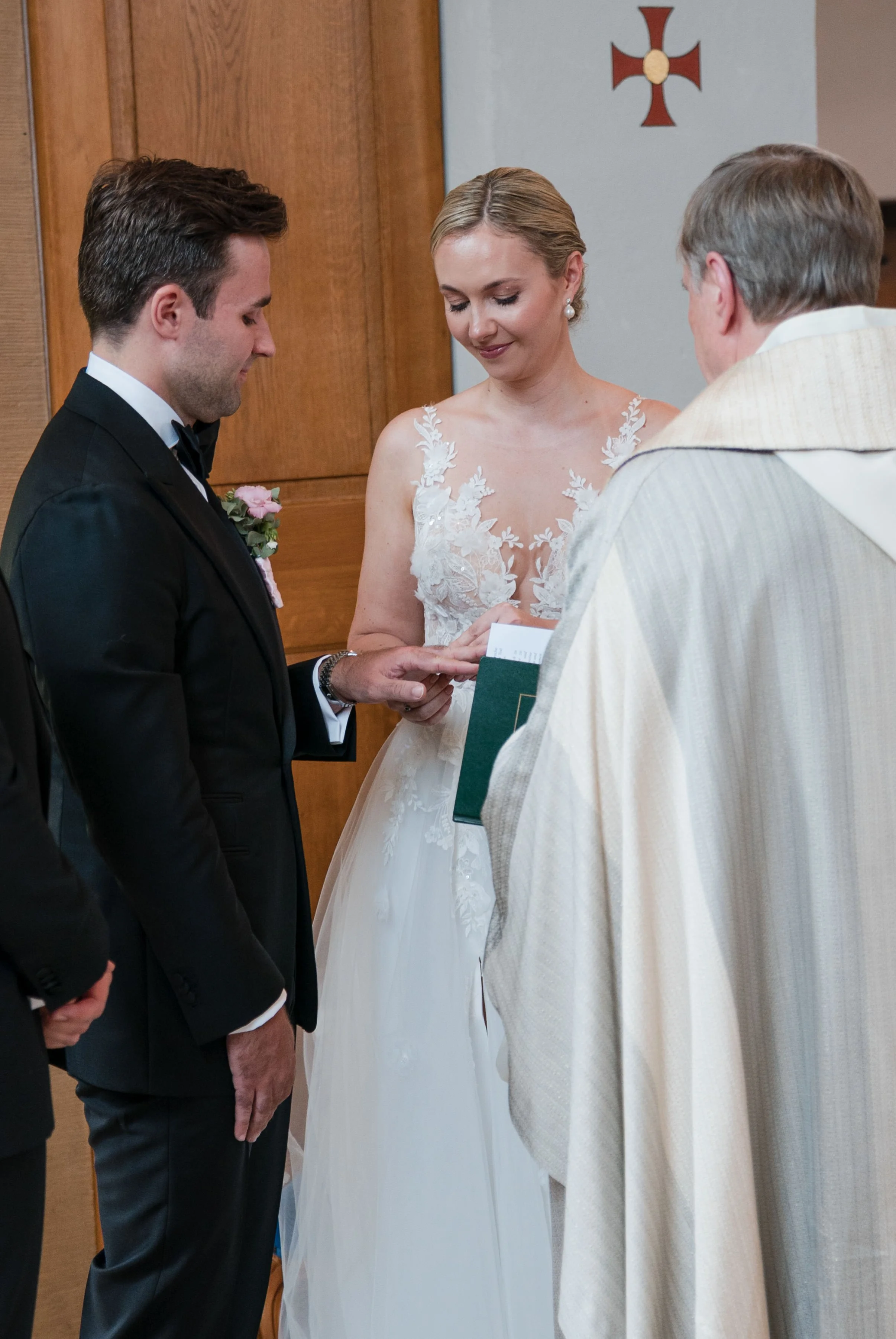 Ein Brautpaar bei der Trauungszeremonie in einer Kirche, mit einem Priester. Die Braut trägt ein weißes Kleid mit Spitzenapplikationen, der Bräutigam ein schwarzes Jackett. Im Hintergrund eine Wand mit einem Kreuz.