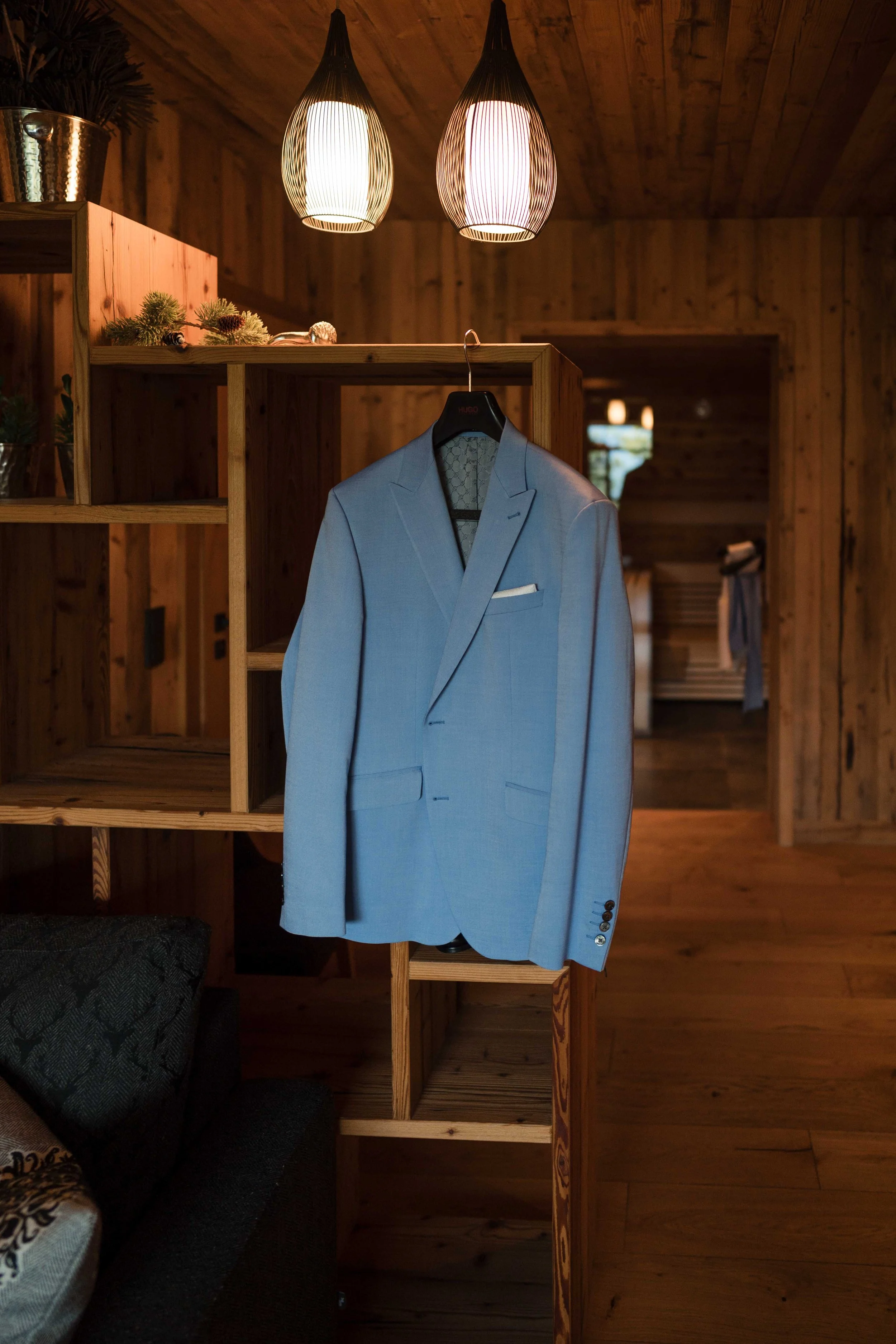 Hellblauer Anzug hängt an einem Haken in einem Holzraum, beleuchtet von zwei modernen Deckenleuchten.
