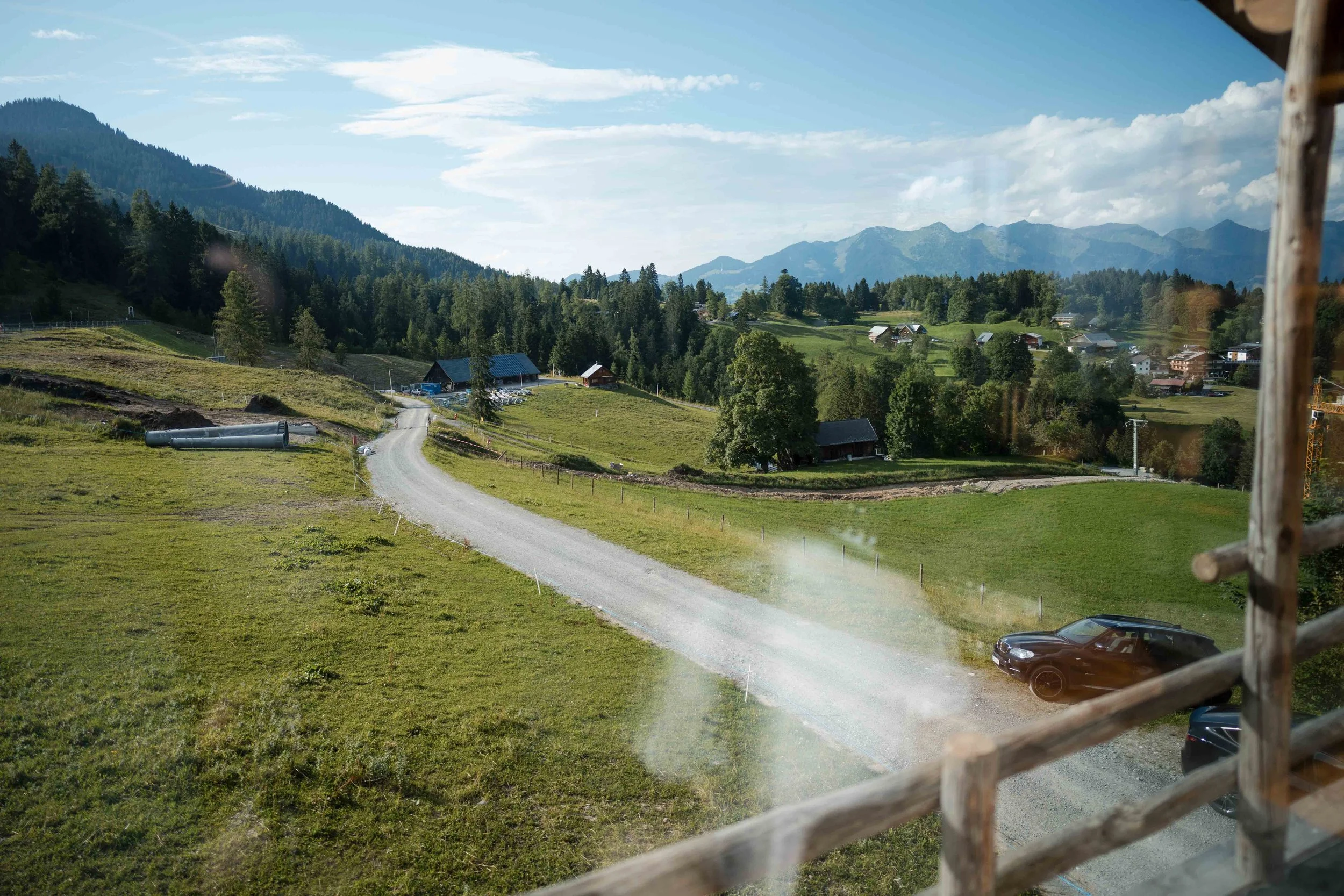 Landschaft mit grünen Wiesen, Bäumen, Hütten und Bergen im Hintergrund, Blick durch ein Fenster mit Holzgittern.