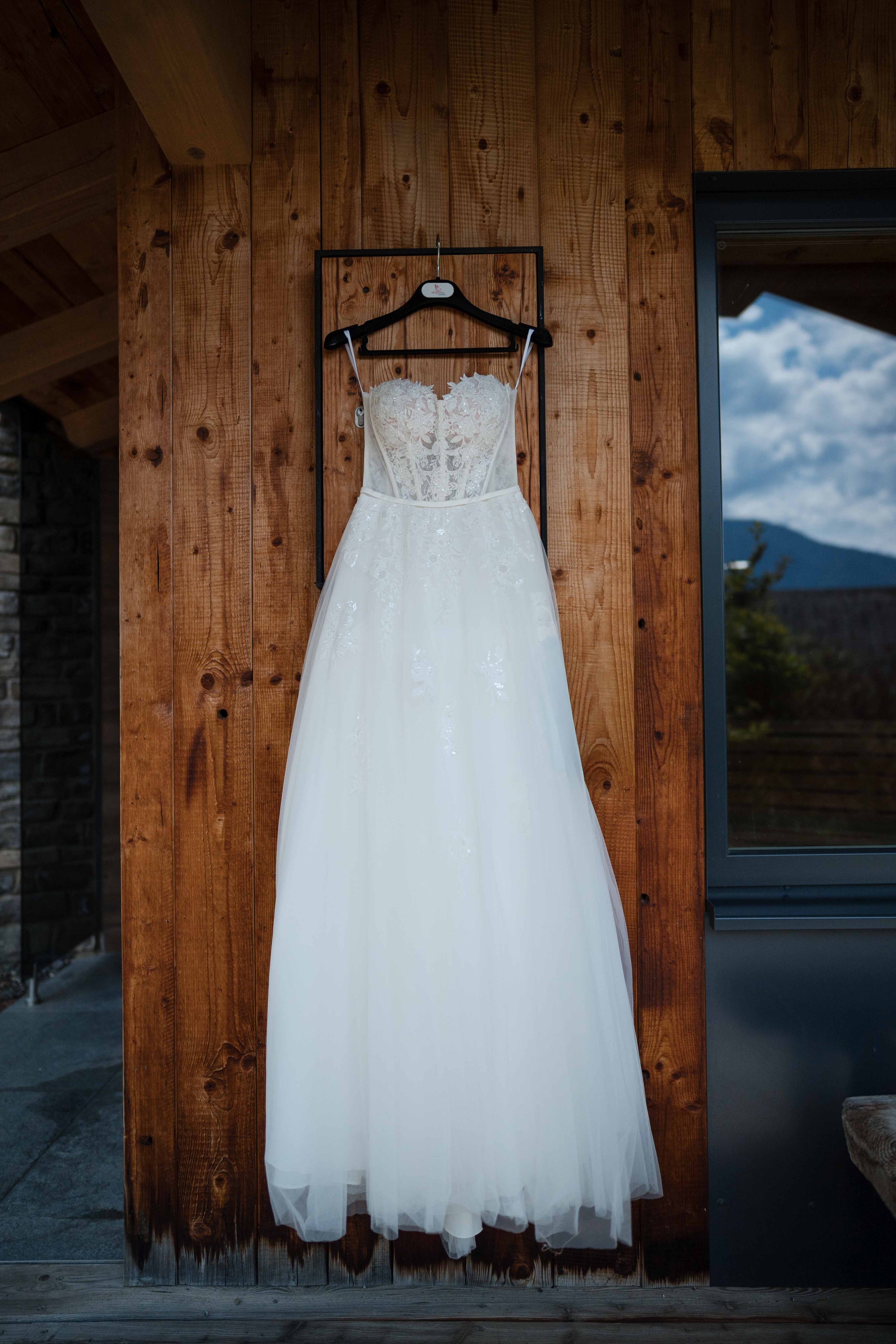 Brautkleid hängt an einer schwarzen Kleiderstange an einer Holzvertäfelung, mit Berg und Himmel im Hintergrund.