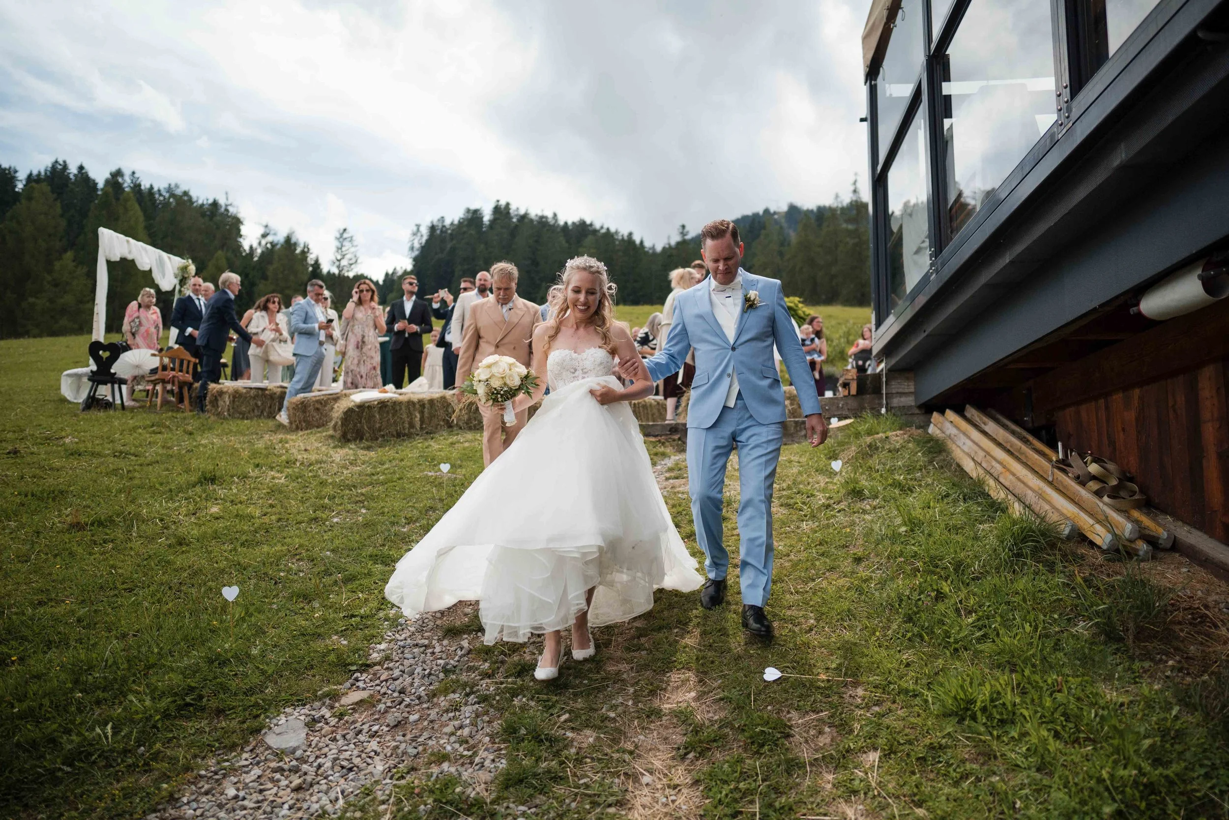 Ein Brautpaar bei ihrer Hochzeit, die braut trägt ein weißes Brautkleid und hält einen Blumenstrauß, der bräutigam trägt ein hellblaues Anzug, während sie gemeinsam auf einer Wiese gehen, im Hintergrund versammelte Gäste bei einer Zeremonie im Freien