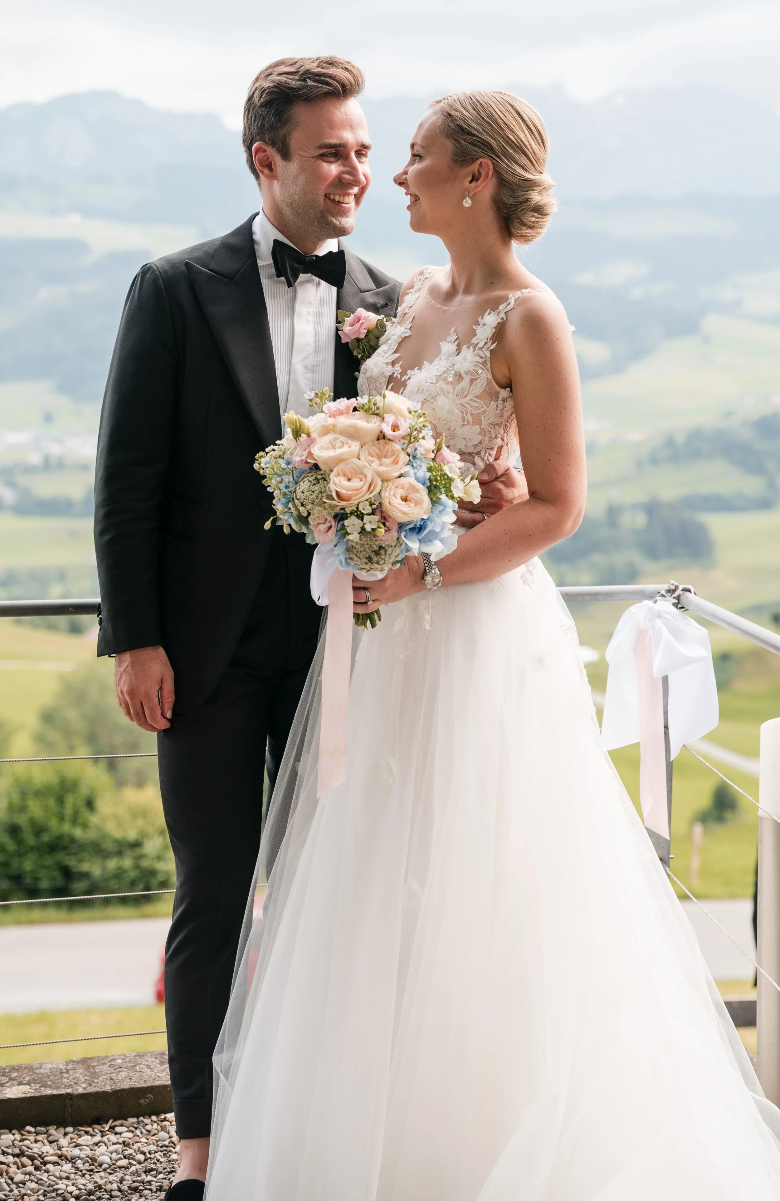 Ein verliebtes Brautpaar bei ihrer Hochzeit, die Braut hält einen Blumenstrauß, im Hintergrund eine ländliche Landschaft.