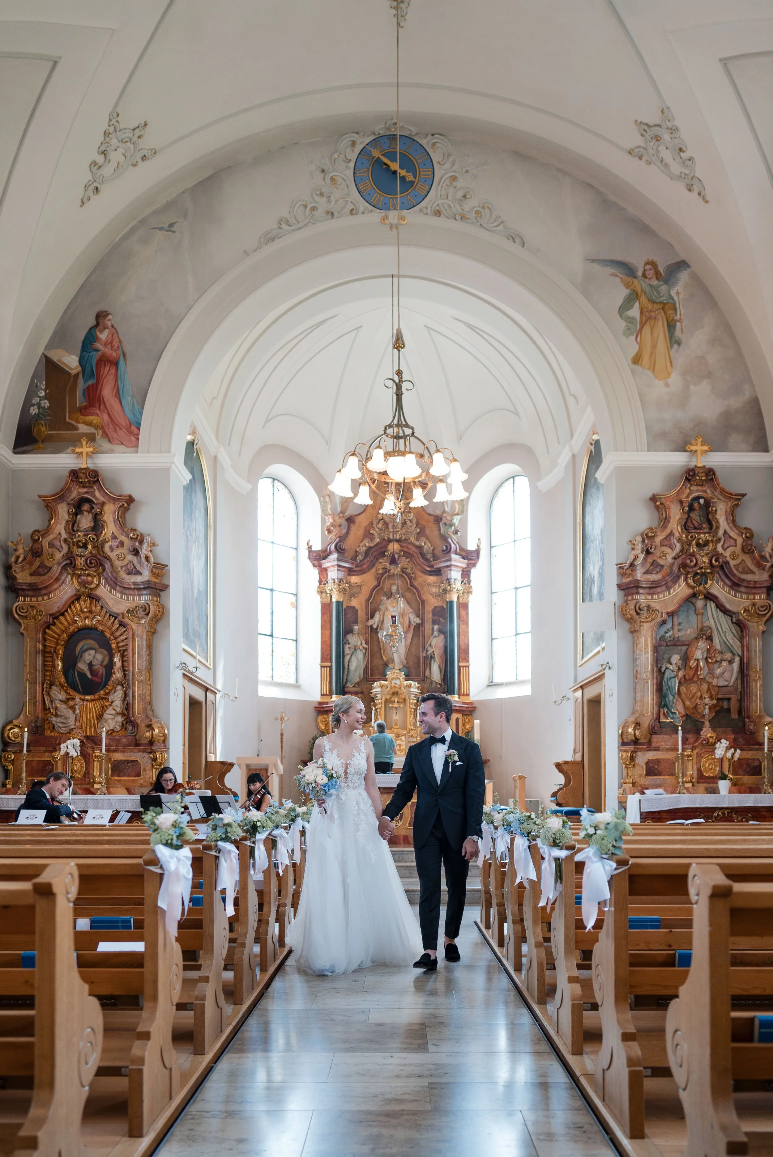 Ein Brautpaar in einer Kirche während ihrer Hochzeitszeremonie, die Braut trägt ein weißes Kleid und hält einen Blumenstrauß, der Bräutigam trägt einen schwarzen Anzug mit Fliege. Im Hintergrund sind Bänke, Kirchenfenster, kunstvolle Altäre und ein K