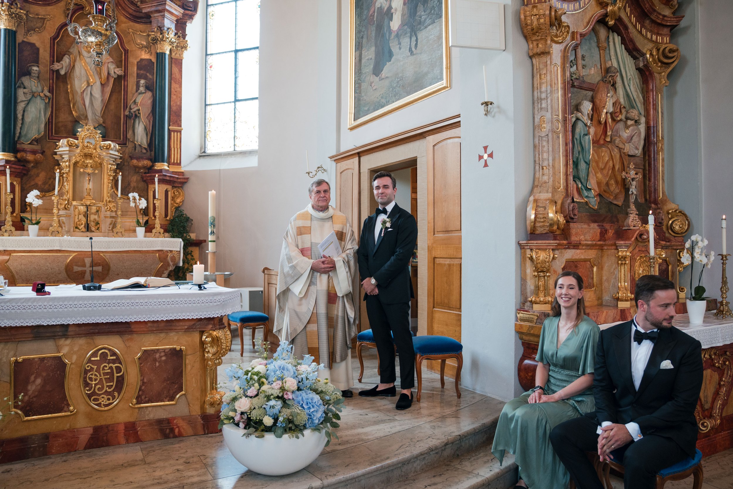 Ein Hochzeit in einer Kirche mit vier Personen, einer Priester und einem Brautpaar, das vor dem Altar steht, sowie eine Frau und einen Mann, die auf Stühlen sitzen.