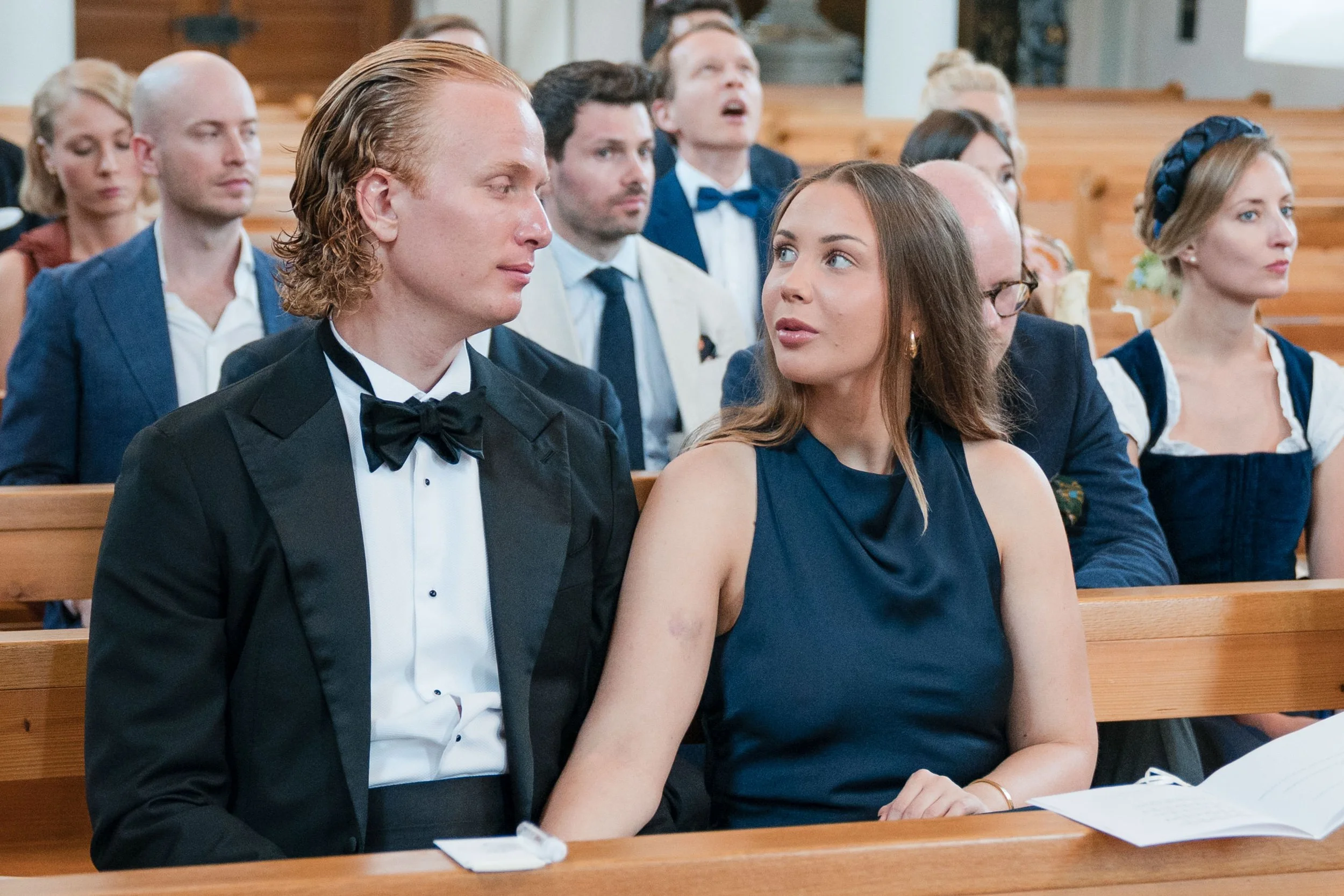 Menschen sitzen in einer Kirche bei einer Hochzeit, der Bräutigam trägt einen schwarzen Anzug mit Fliege, die Braut trägt ein dunkelblaues Kleid, beide schauen sich an.