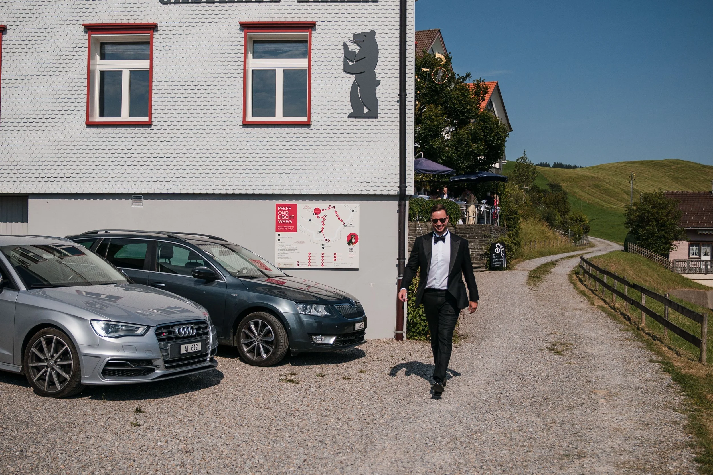 Mann in Smoking läuft auf einem Kiesweg an geparkten Autos vorbei, im Hintergrund ein Gebäude mit einem Bärenkunstwerk an der Fassade und einer Terrasse mit Gästen, umgeben von grünen Hügeln und Bäumen.