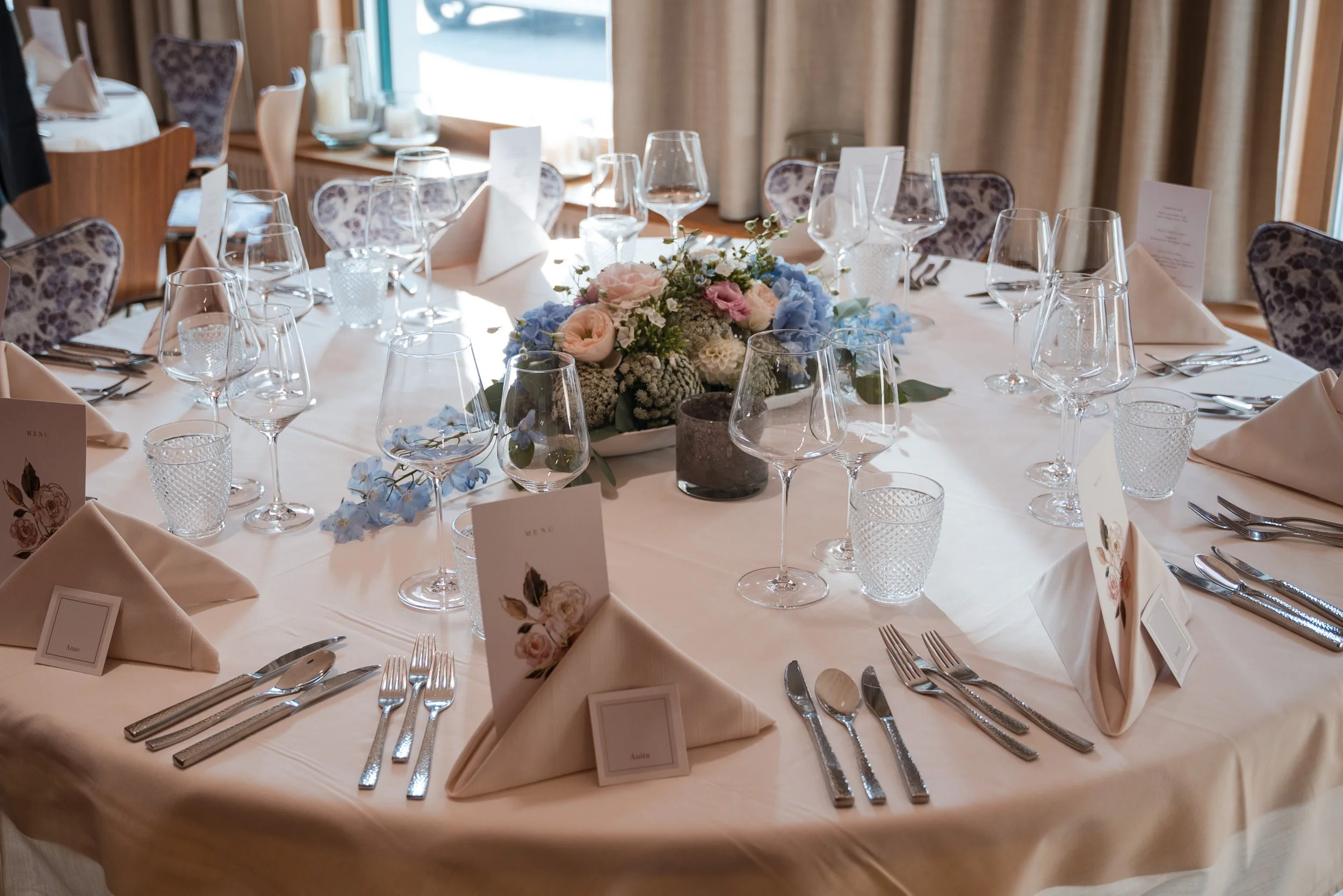Ein schön gedeckter runder Esstisch mit Blumenstrauß in der Mitte, mehreren Weingläsern, Wasserbechern, Besteck, weißen Stoffservietten und Menü-Karten in einem festlichen Raum mit natürlichem Licht.