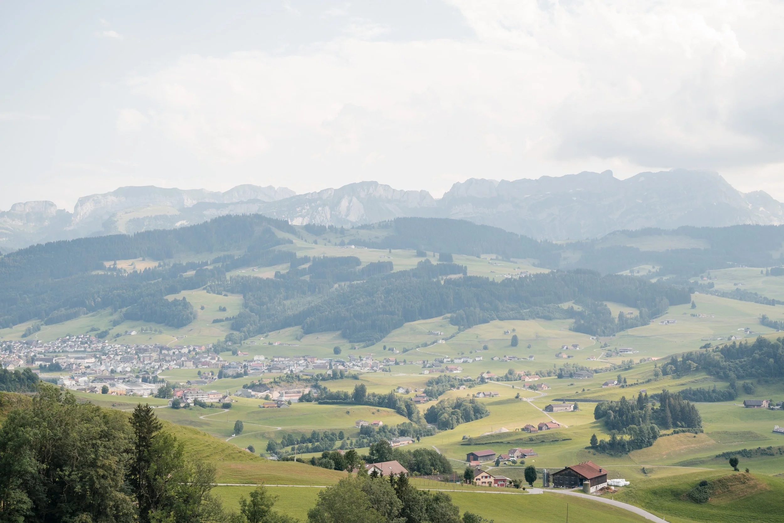 Landschaft mit Hügeln, Feldern, Häusern und Bergen im Hintergrund.