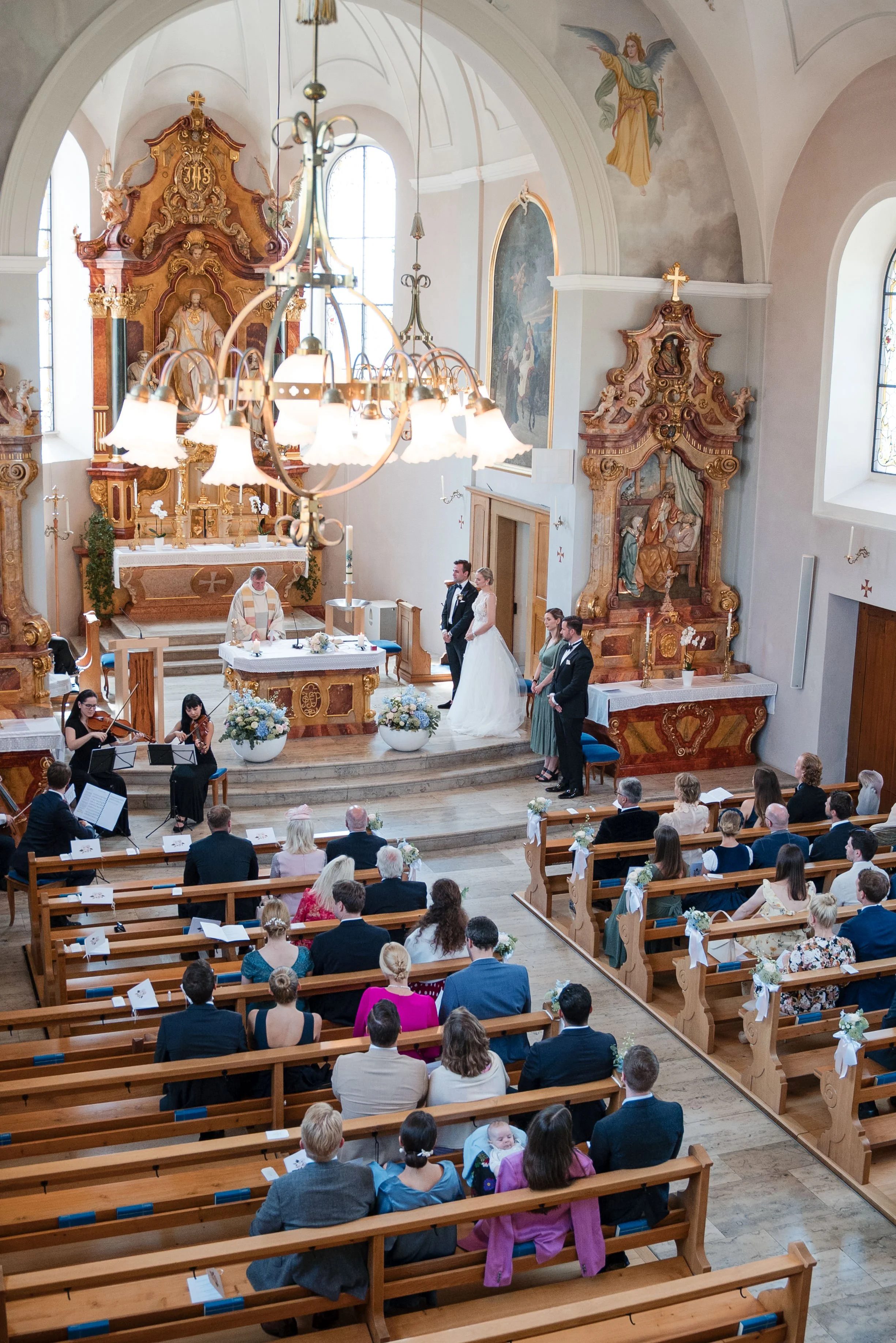 Ein Hochzeitsgottesdienst in einer Kirche mit einem Priester am Altar, Braut und Bräutigam vor dem Altar, Gäste auf Bänken, klassische Dekorationen, Kirchenfenster und religiöse Kunst an den Wänden.