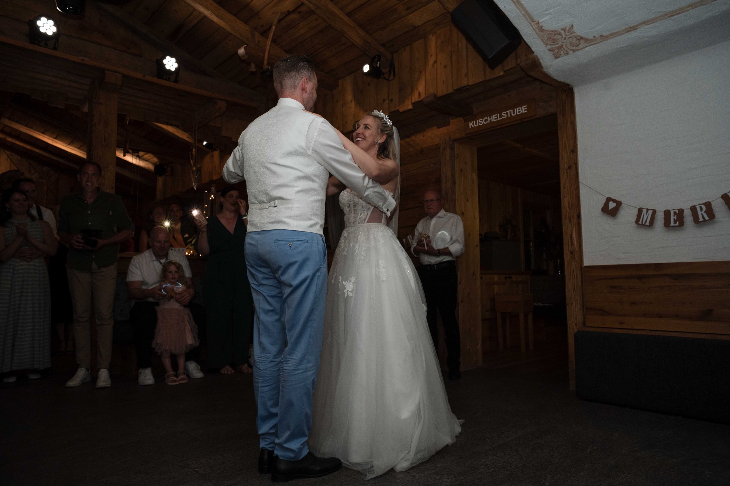 Braut und Bräutigam tanzen auf der Hochzeitsfeier, umgeben von Gästen, im Holzraum mit Dekorationen und einem Banner mit Herzen.