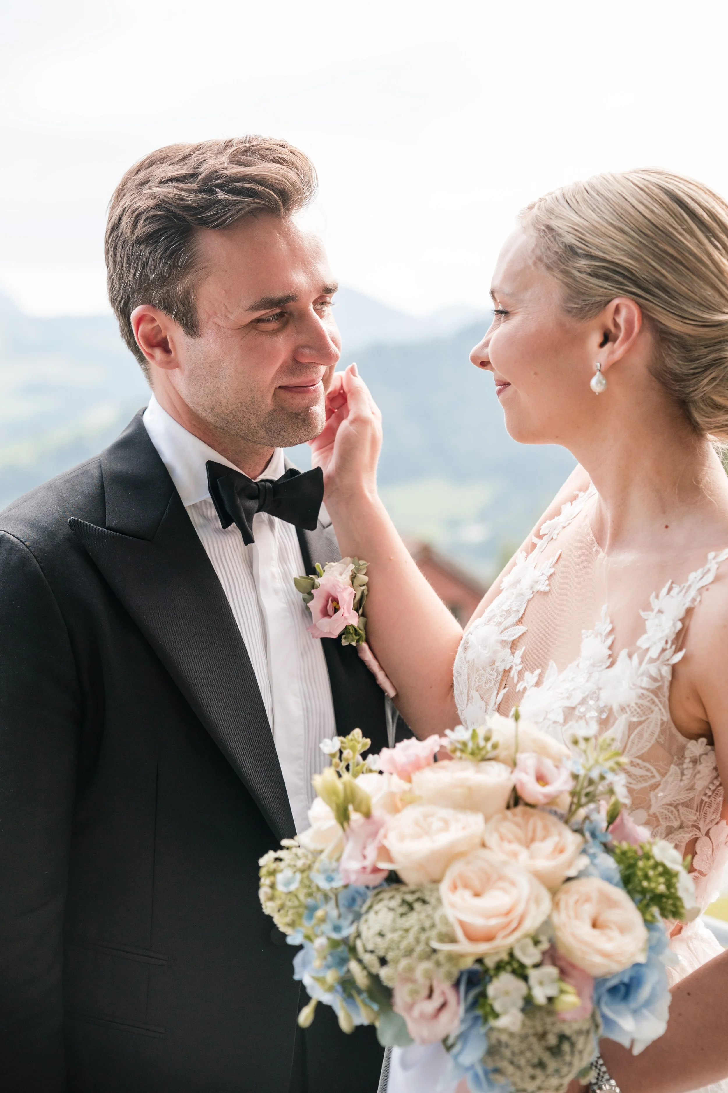 Ein Brautpaar, das sich liebevoll ansieht, bei einer Hochzeitszeremonie im Freien, mit Bergen im Hintergrund, die Braut hält einen Blumenstrauß.