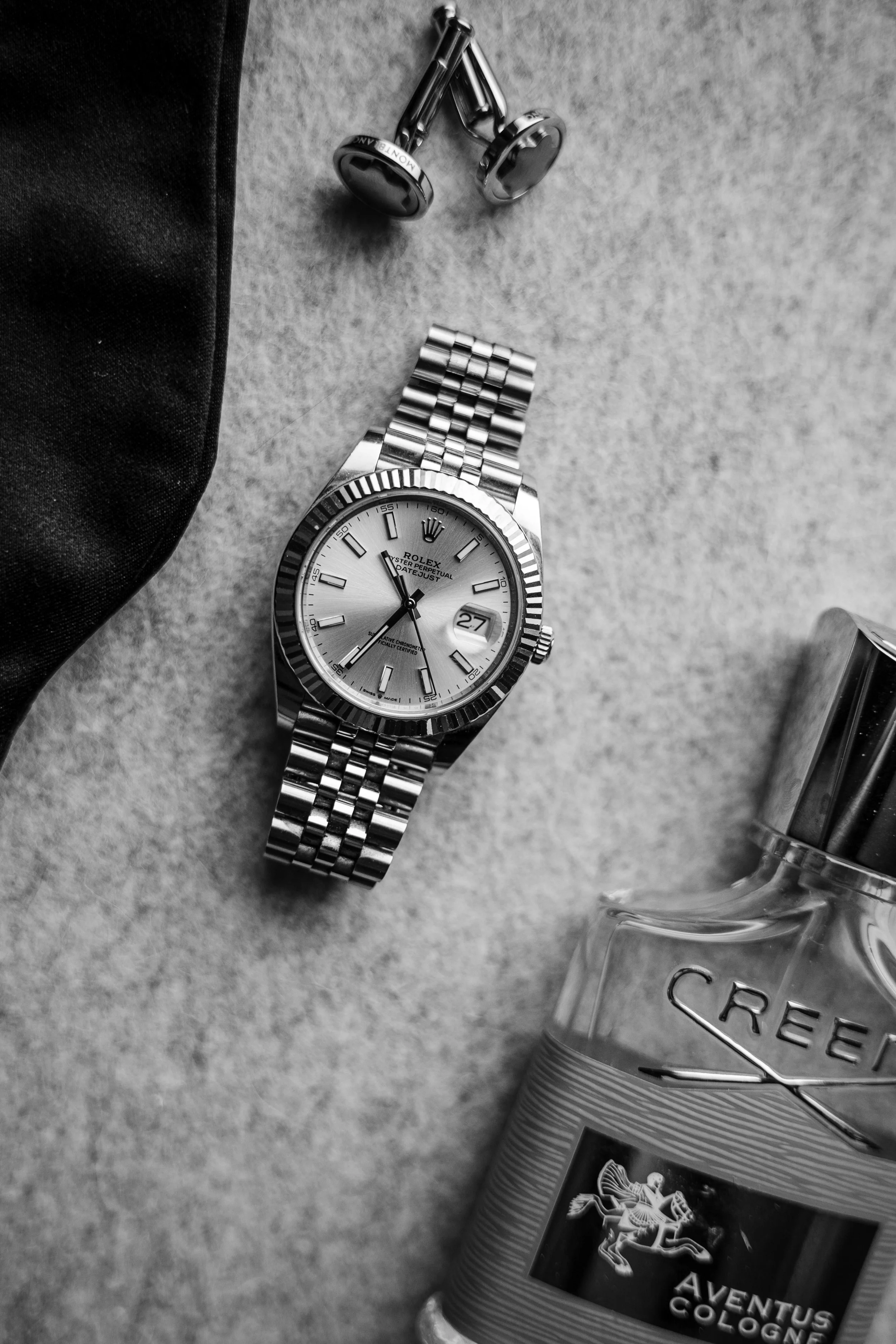 Schwarz-weiße Nahaufnahme eines Rolex Armbanduhr, ein Parfümflasche, eine Kappe, und Ohrhörer auf einem grauen Stoffhintergrund.