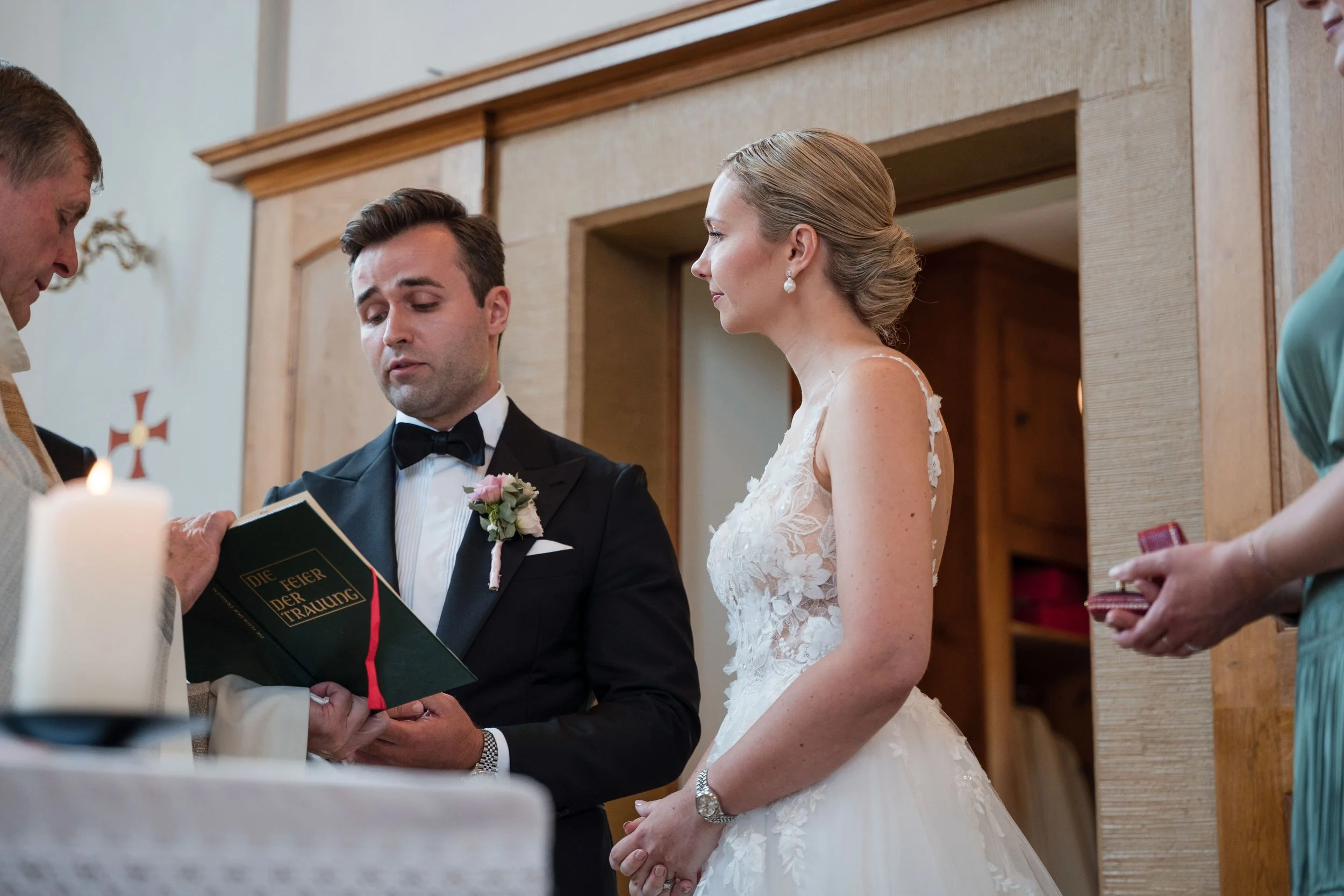 Ein Brautpaar bei der Trauung steht vor einem Priester, der ein Buch mit dem Titel 'Die Feier der Trauung' hält. Der Bräutigam trägt einen schwarzen Smoking mit Fliege und eine rosa Blume im Revers. Die Braut trägt ein weißes Hochzeitskleid mit Spitz