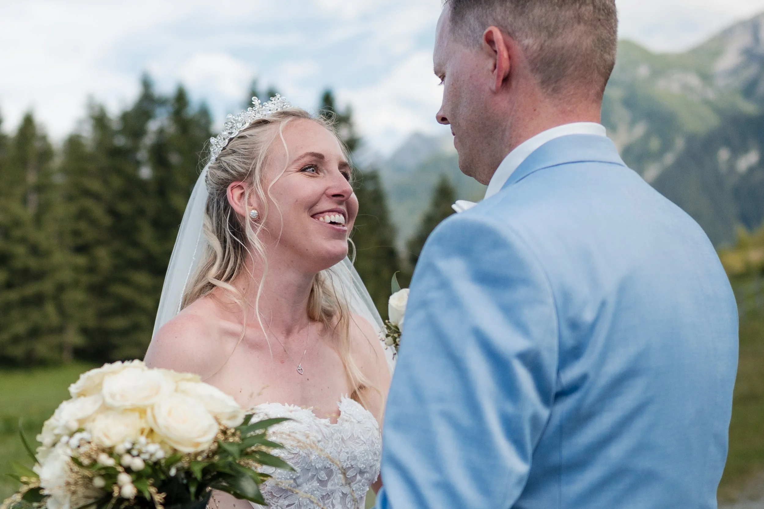 Braut und Bräutigam bei ihrer Hochzeit im Freien mit Bergen im Hintergrund, die Braut hält einen Blumenstrauß, beide lächeln sich verliebt an.