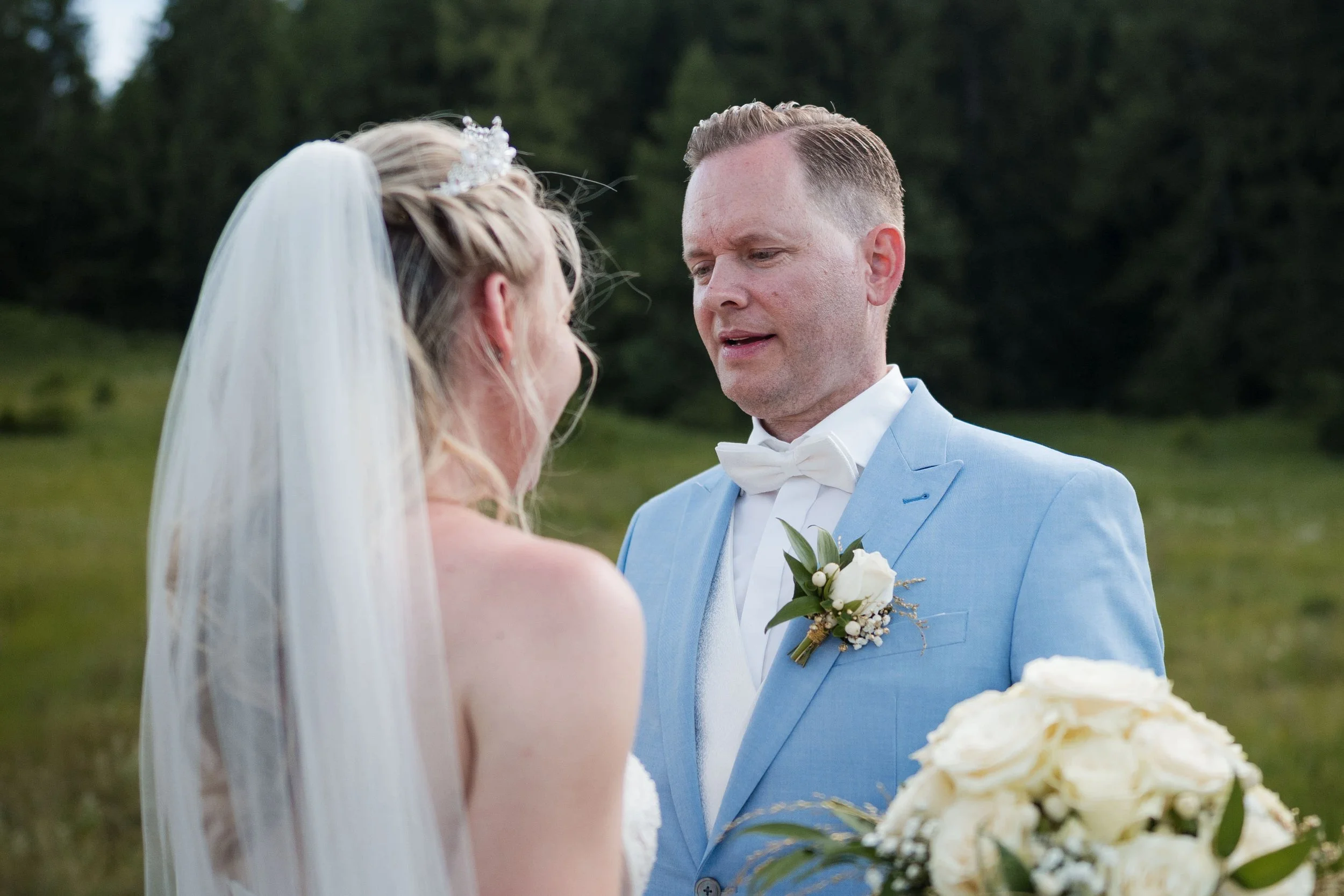 Ein Brautpaar im Freien bei einer Hochzeit, der Bräutigam trägt einen hellblauen Anzug mit Fliege, die Braut trägt ein Hochzeitskleid mit Schleier und eine Tiara, sie steht sich gegenüber, im Hintergrund sind Bäume und Gras zu sehen.