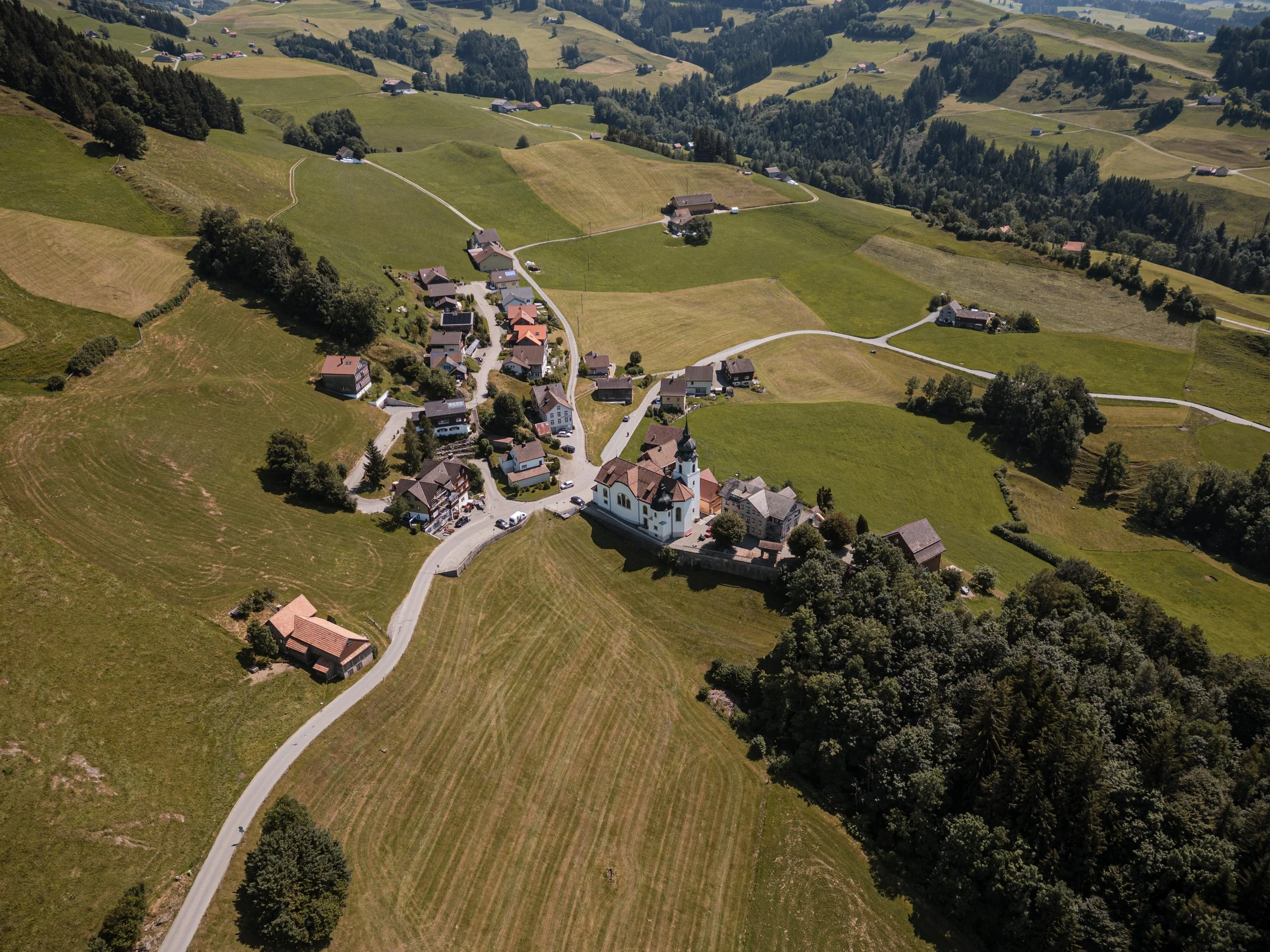Ein Luftbild eines kleinen, ländlichen Dorfes auf einer hügeligen, grün bewachsenen Landschaft mit Feldern und Wäldern.