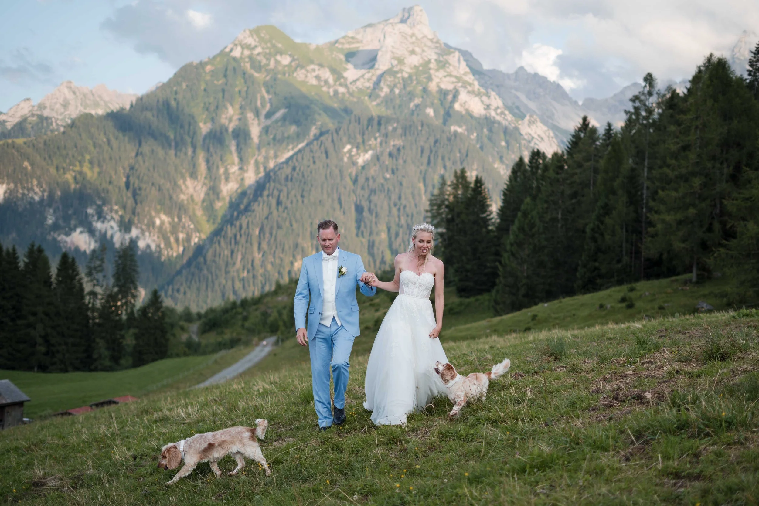 Ein Brautpaar in Hochzeitskleidung läuft auf einer grünen Wiese mit Hunden vor einer Bergkulisse in den Alpen.
