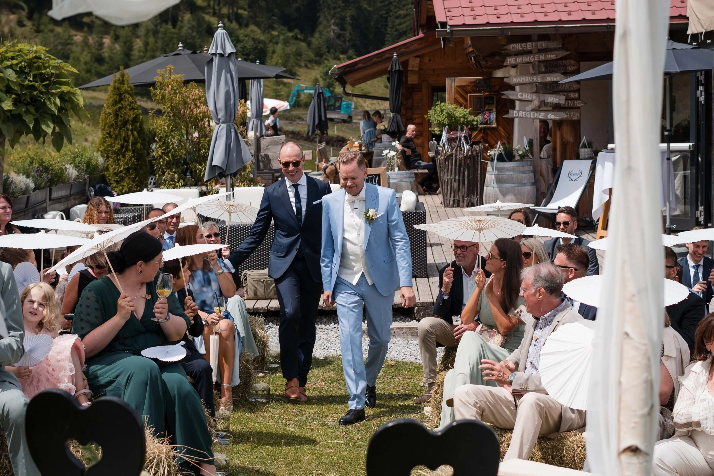 Ein Hochzeitsfest im Freien mit einem Paar in Hellblauem Anzug, umgeben von Gästen mit Sonnenschirmen, in einer ländlichen Umgebung mit Bergen und Wäldern im Hintergrund.