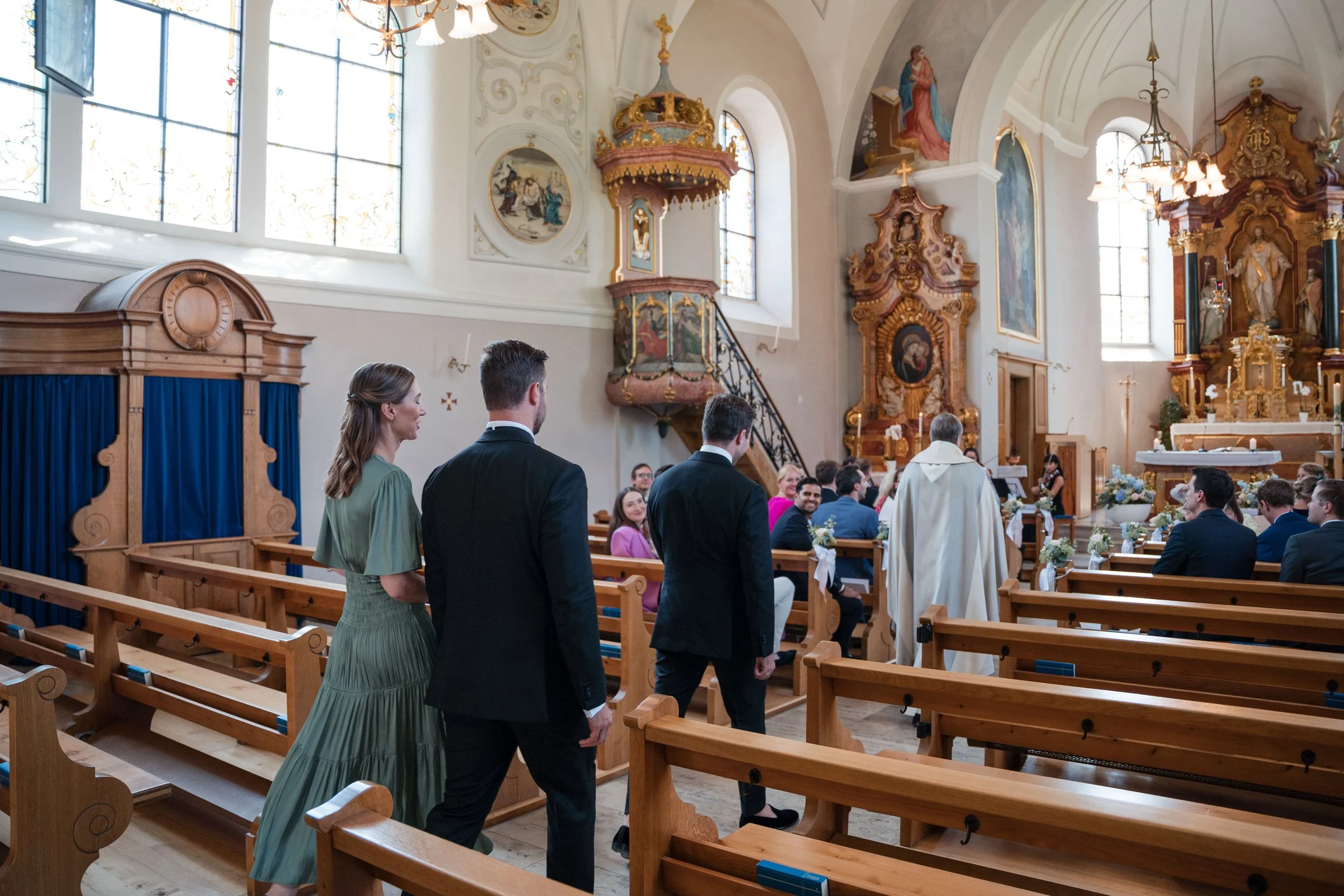 Paare sitzen in einer Kirche während einer Hochzeitszeremonie, Priester in weißen Gewand steht vorne, alte Holzbankreihen und bunte Blumen auf den Bänken.