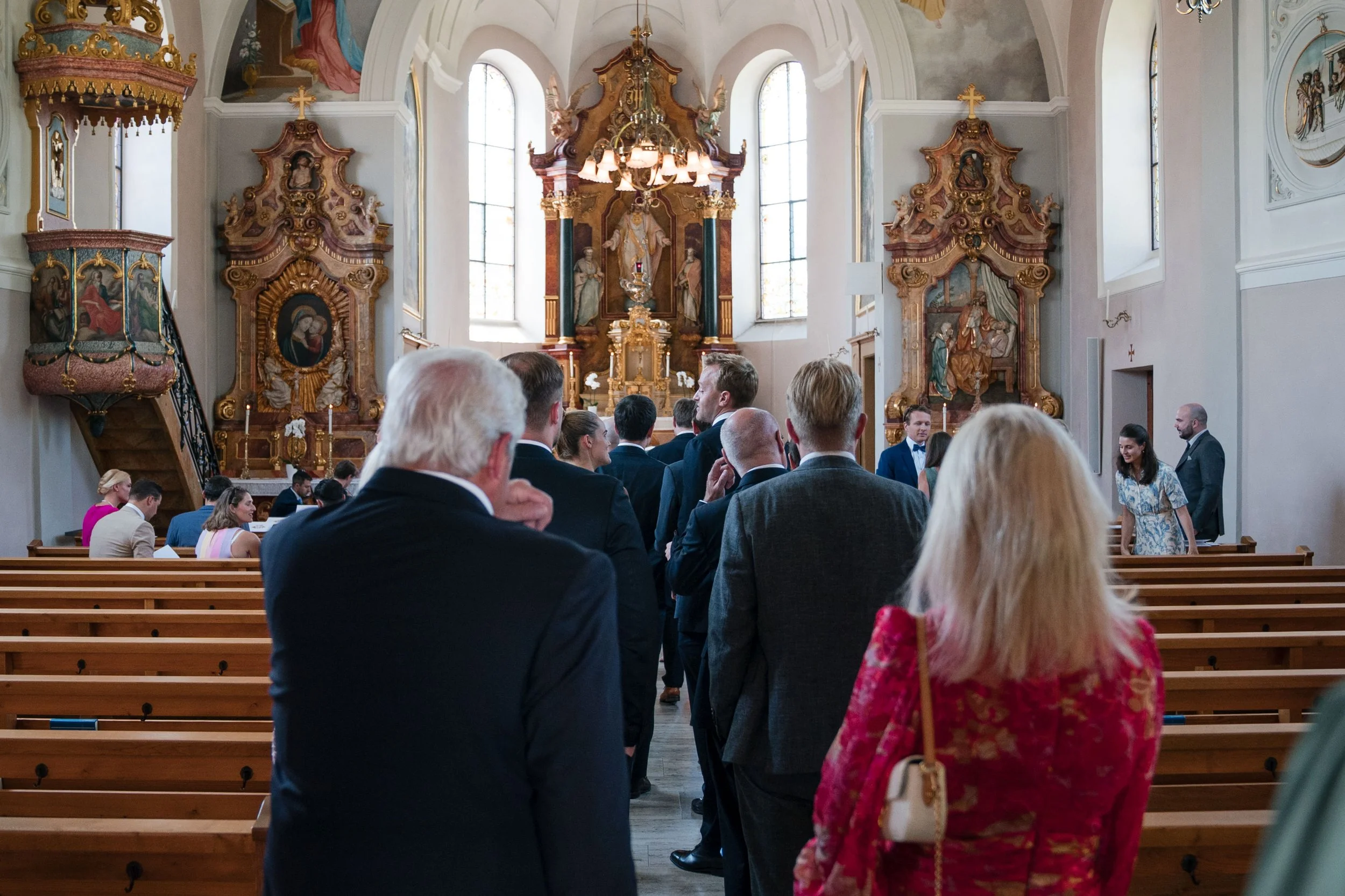 Menschen in formeller Kleidung, die in einer Kirche anstehen, vermutlich für eine Zeremonie wie eine Hochzeit, mit Barock-Altargestaltung im Hintergrund.