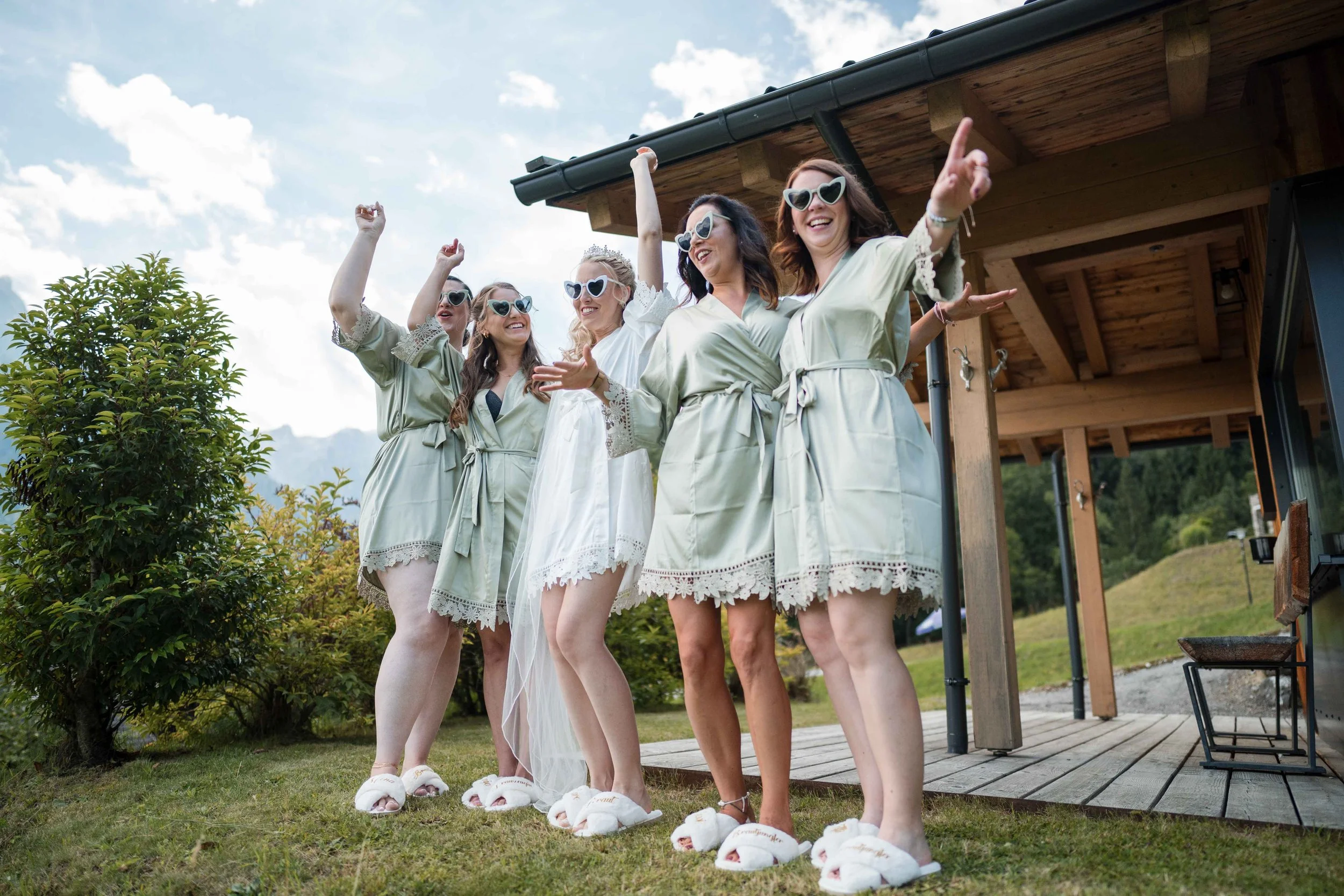 Fünf Frauen in Bademänteln und mit Herz-Sonnenbrillen feiern draußen auf einer Veranda, einen Junggesellinnenabschied, lachend und mit Armen in die Luft gestreckt.