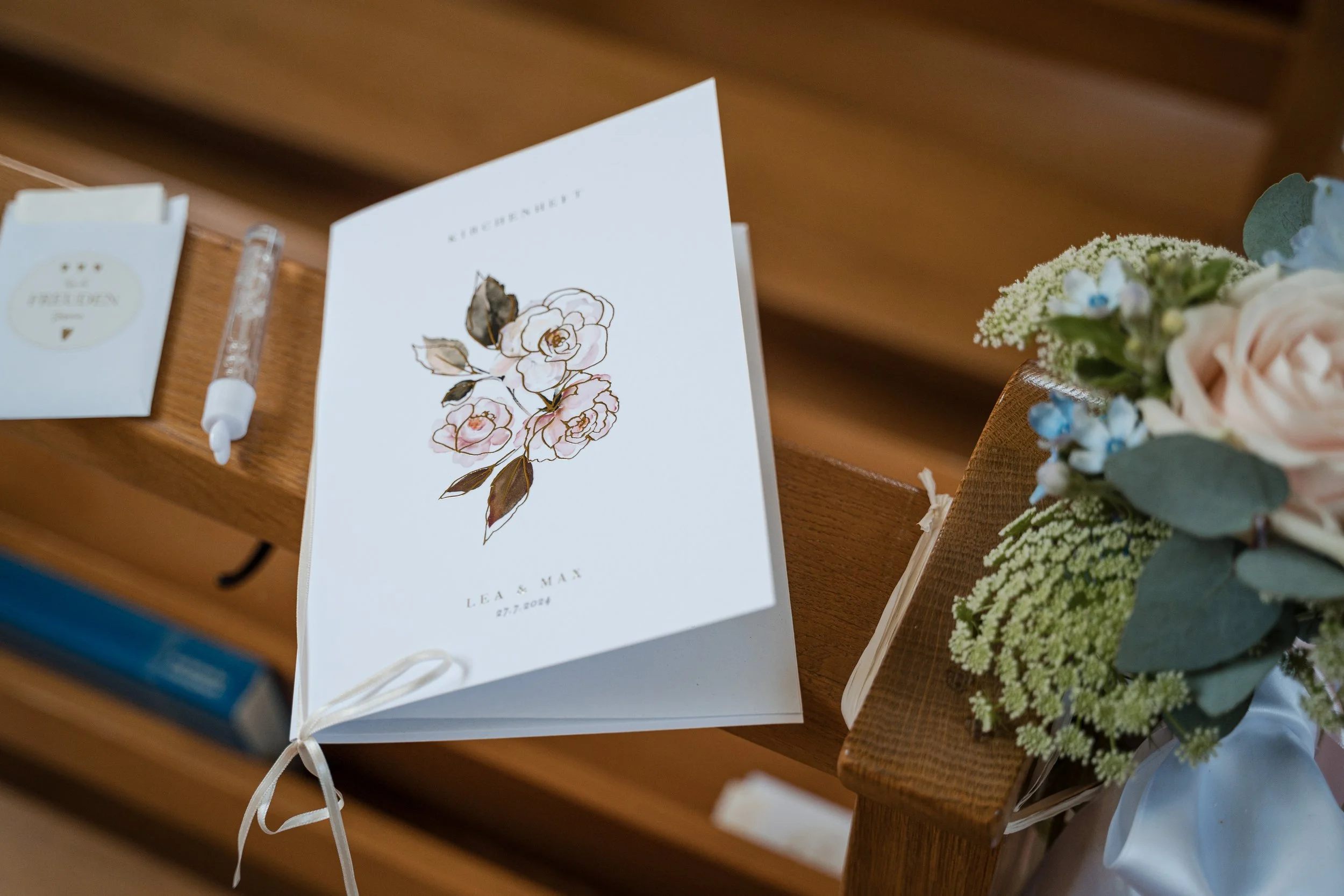 Ein Hochzeitsgästebuch mit einer Blumenzeichnung liegt auf einer Holzbank, daneben befindet sich ein Stift, und auf der rechten Seite eine Blumenstrauß aus Rosé, weißen und blauen Blumen mit grünem Laub.