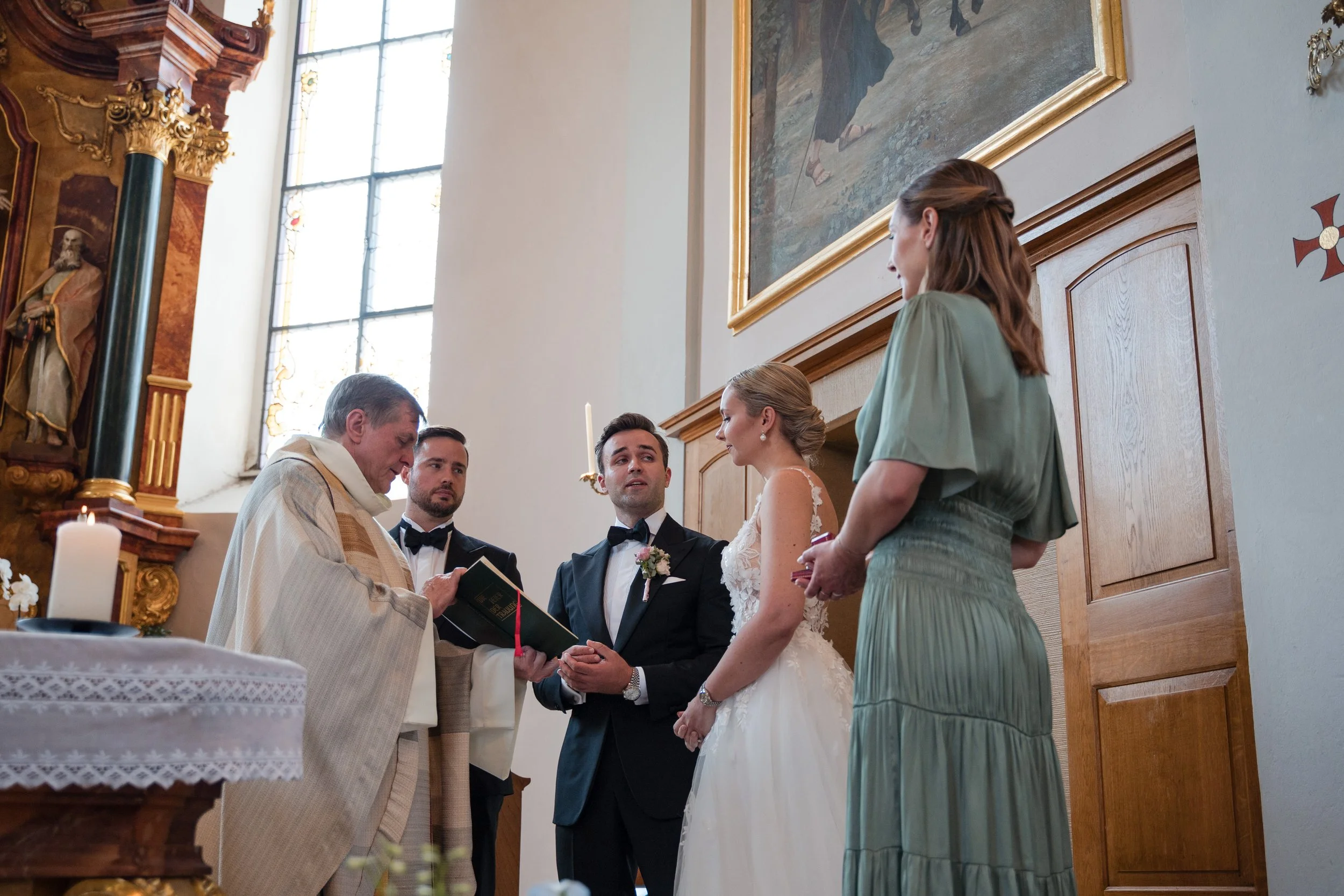 Ein Brautpaar steht während einer kirchlichen Zeremonie vor einem Priester. Der Bräutigam trägt einen schwarzen Anzug mit Fliege, die Braut ein weißes Hochzeitskleid. Zwei Gäste, eine Frau in einem grünen Kleid und ein Mann in einem schwarzen Anzug, 