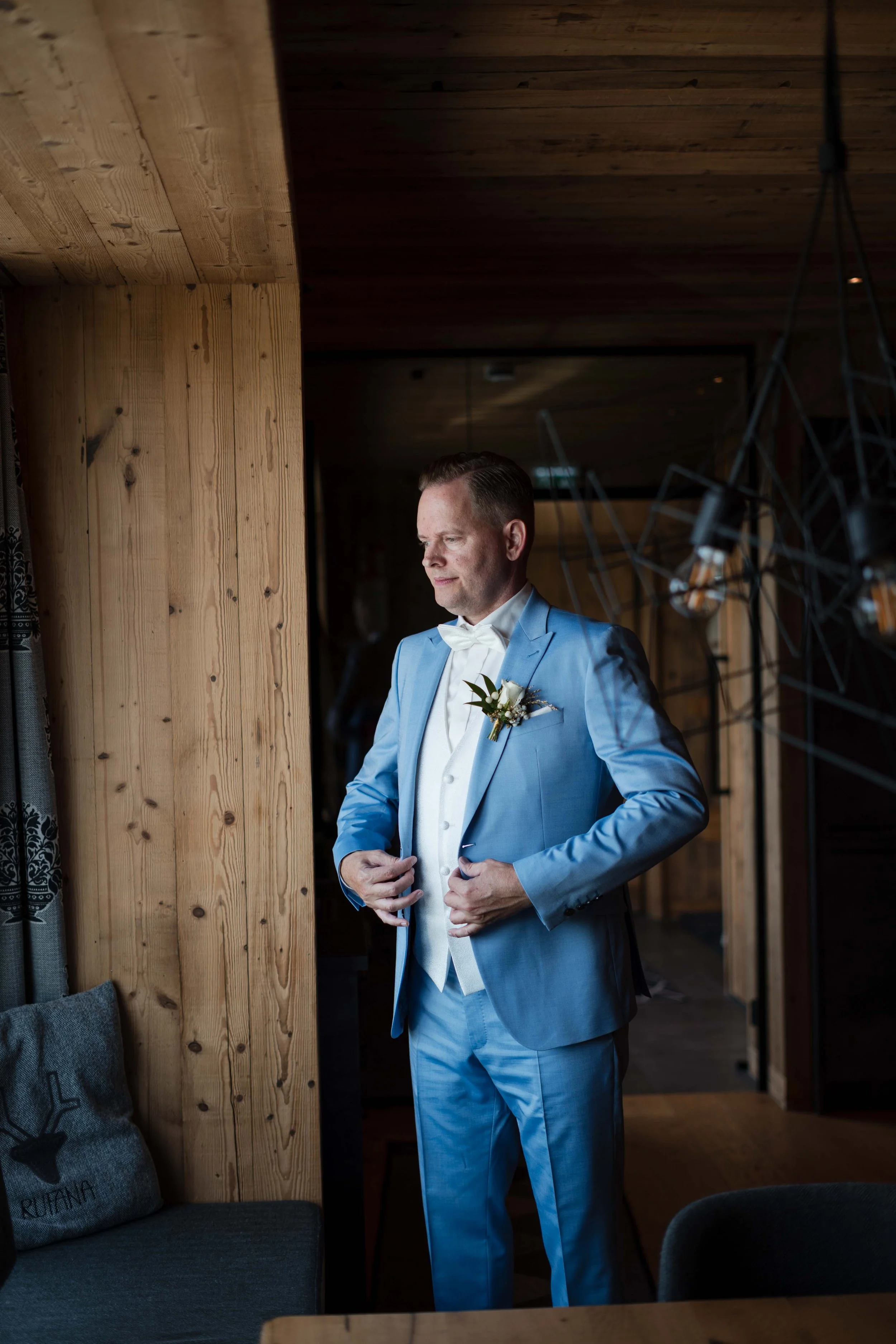Ein Mann in helblauem Anzug mit Fliege steht vor einem Fenster im Raum, offensichtlich an seinem Hochzeitstag.