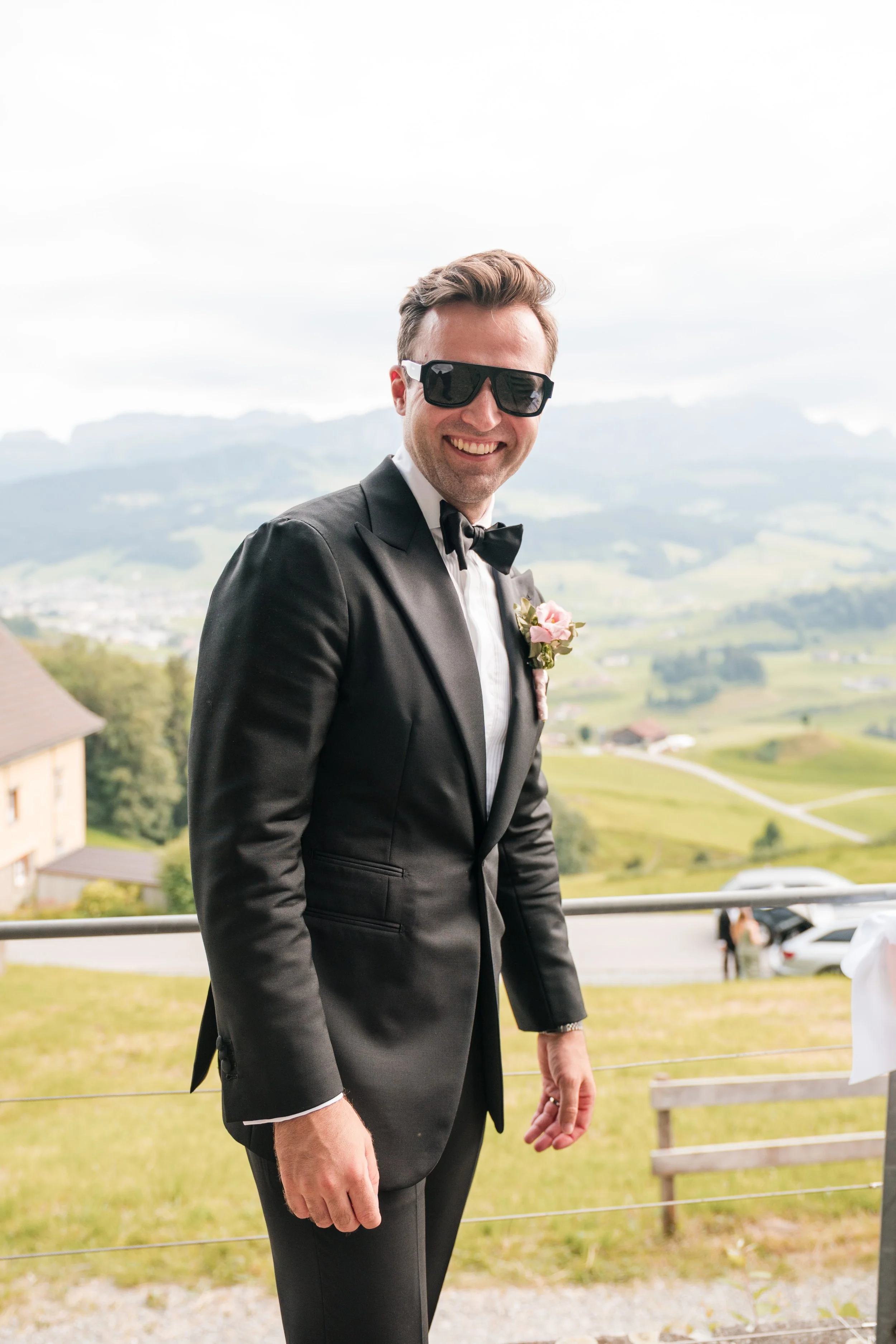 Mann in schwarzem Anzug, Sonnenbrille und Fliege beim Hochzeitsfoto im Freien mit Hügeln und Häuser im Hintergrund.