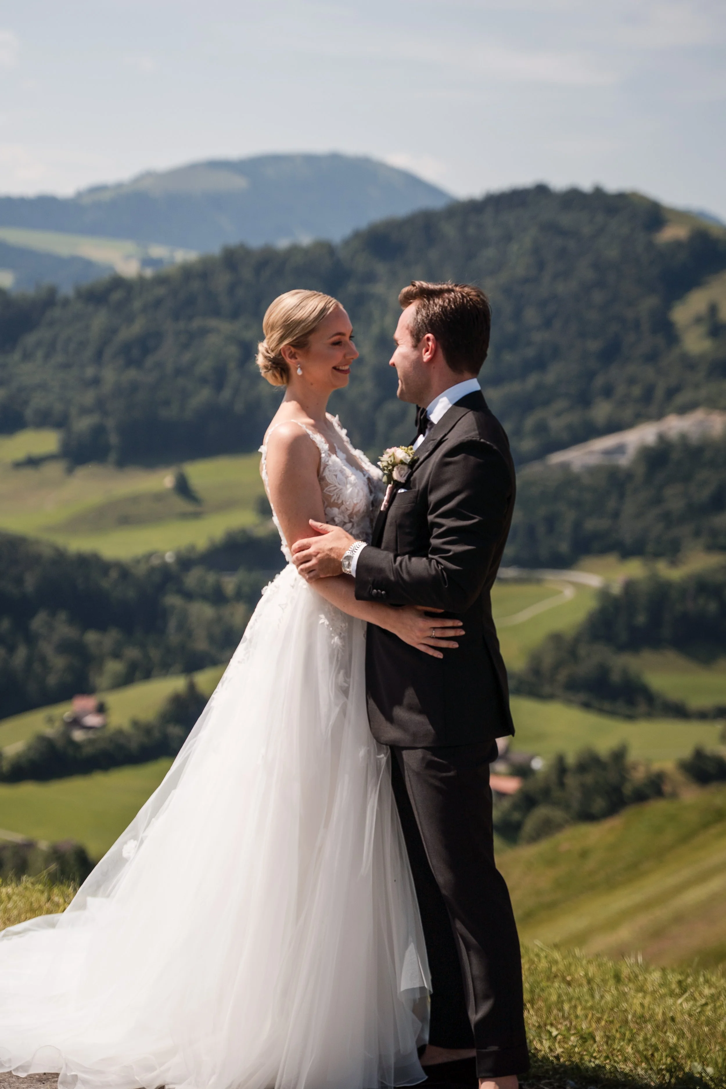 Ein Brautpaar bei einer Hochzeit im Grünen, vor einer hügeligen Landschaft mit Bäumen und Bergen im Hintergrund.