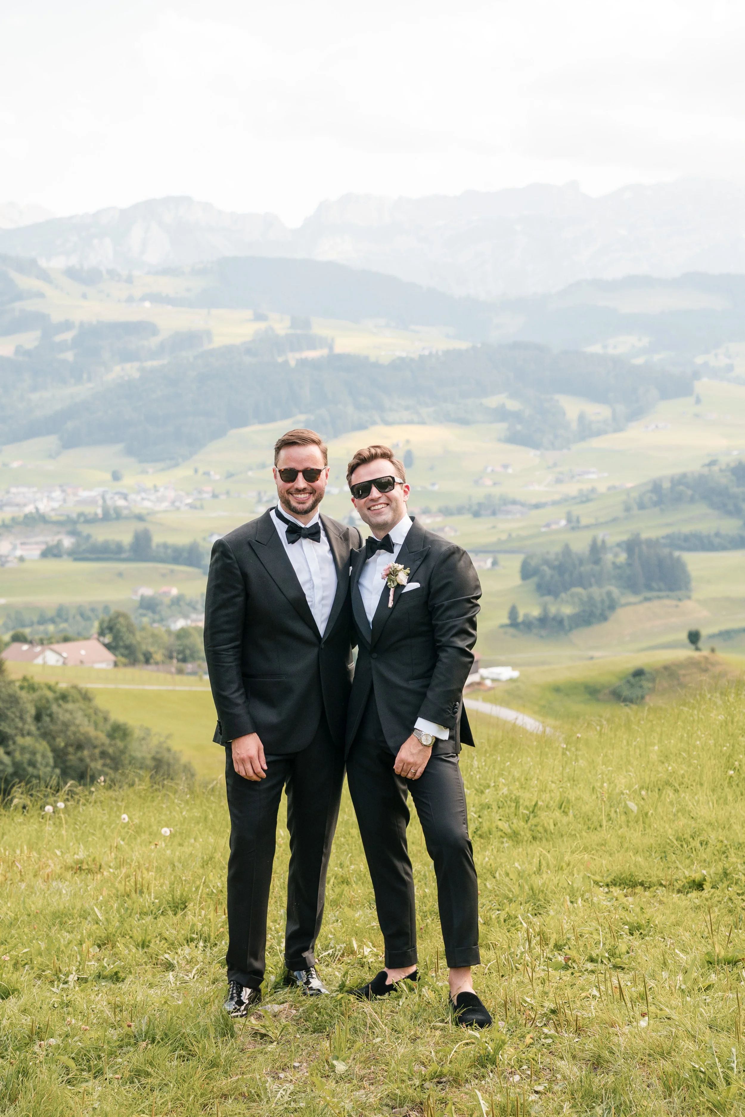 Zwei Männer in Smokings stehen in einer grünen Wiese vor einer bergigen Landschaft, beide tragen Sonnenbrillen und lächeln.