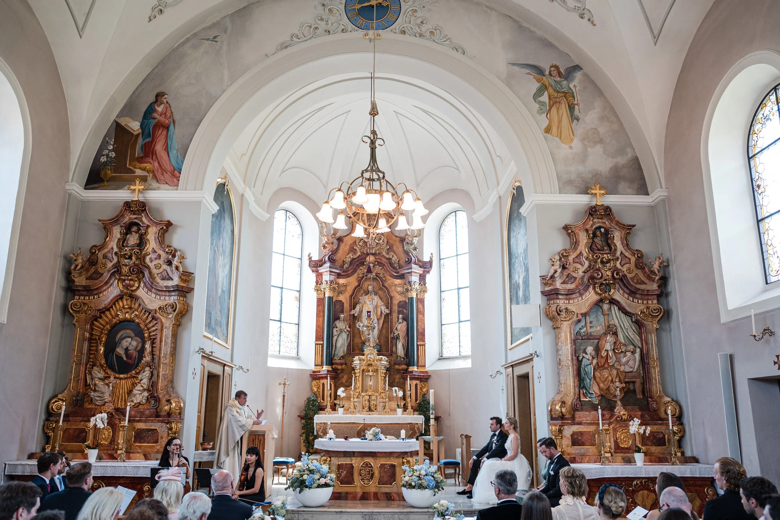 Eine Hochzeitszeremonie in einer Kirche, bei der ein Priester vor einem Altar steht. Zwei Personen in formeller Kleidung, scheinbar Braut und Bräutigam, sitzen auf der rechten Seite. Die Kirche ist reich verziert mit barockartigen Holzvertäfelungen, 